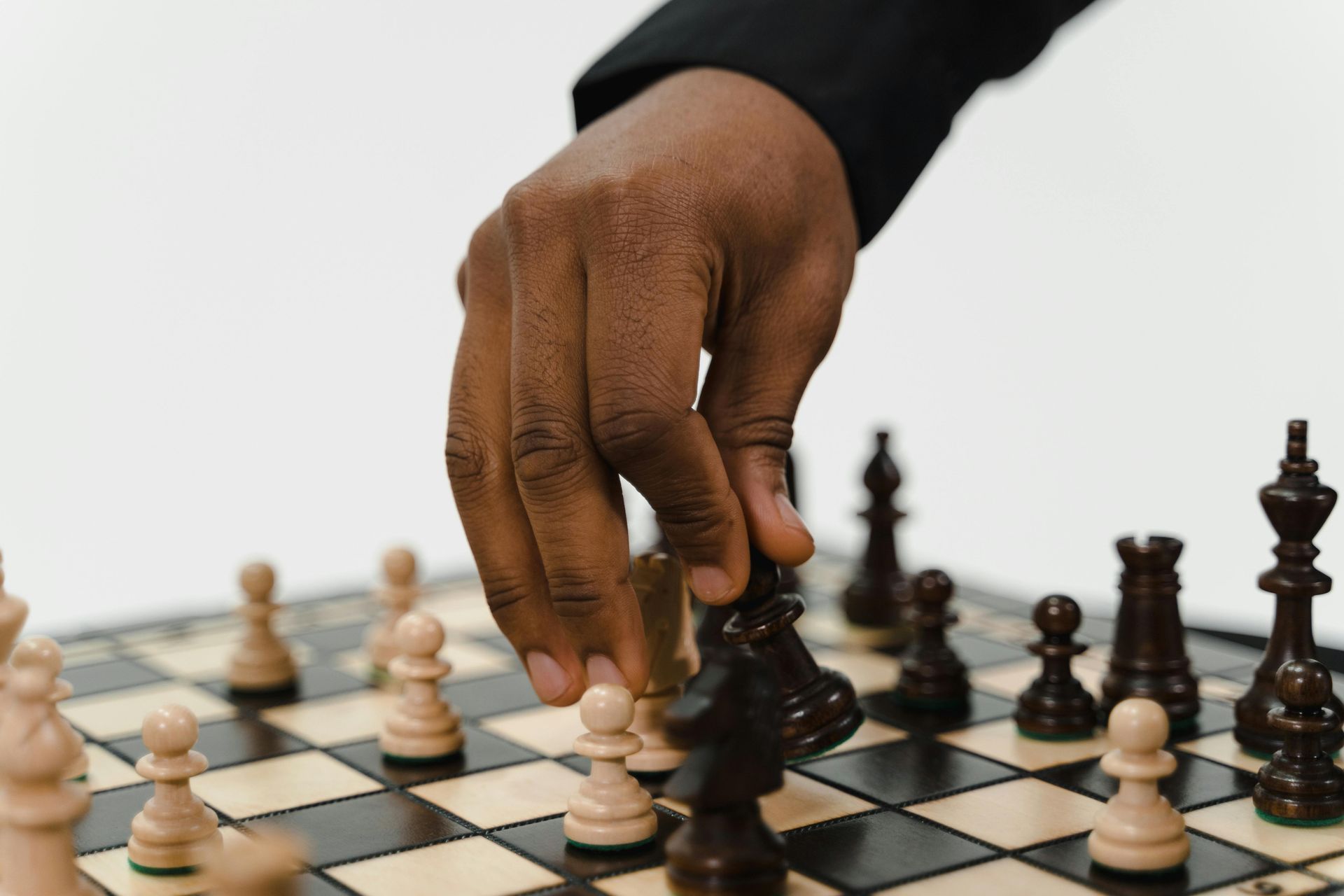 A dark-skinned hand moves a black chess piece on a chessboard with black and white pieces, against a white background.