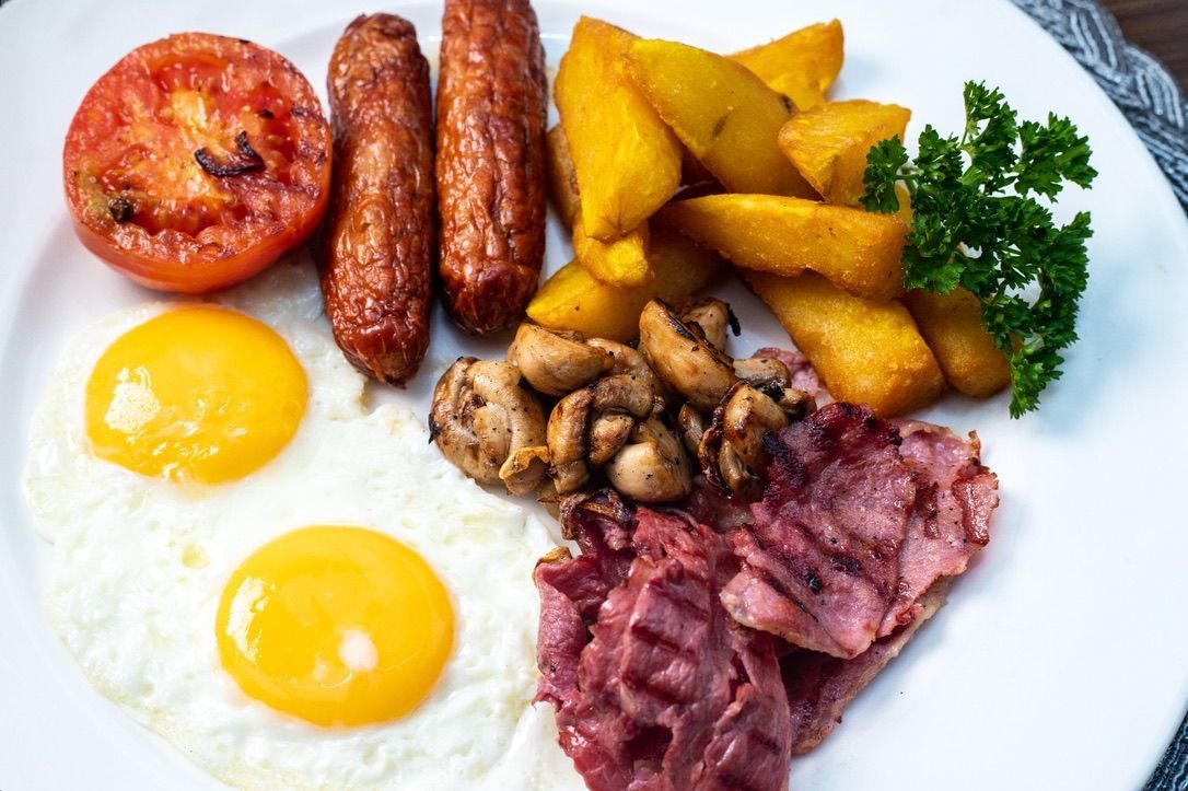 A full English breakfast on a white plate: fried eggs, sausage, bacon, potatoes, tomato, and mushrooms.