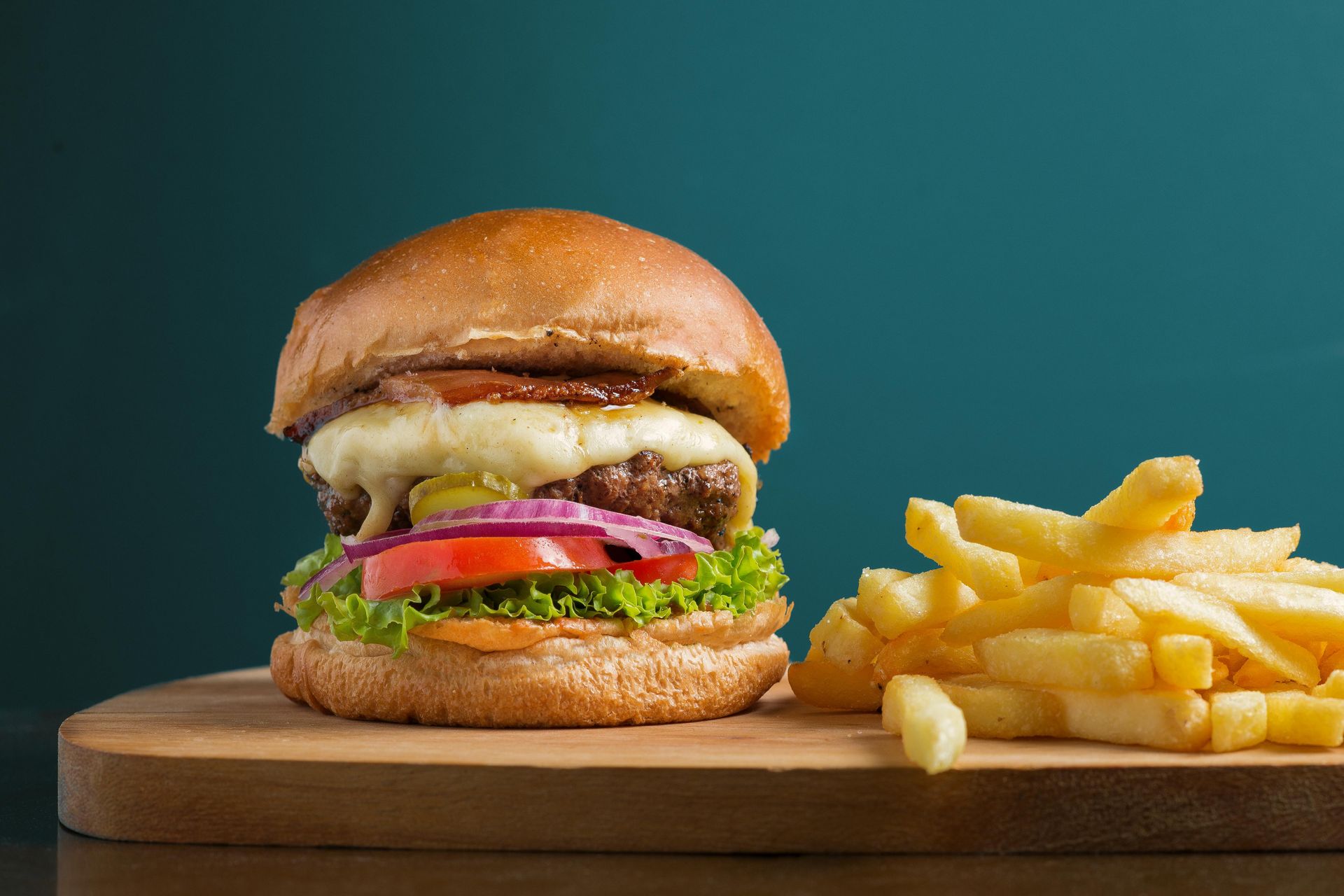 Burger with cheese, red onion, tomato, and lettuce on a bun, served with french fries on a wooden board.