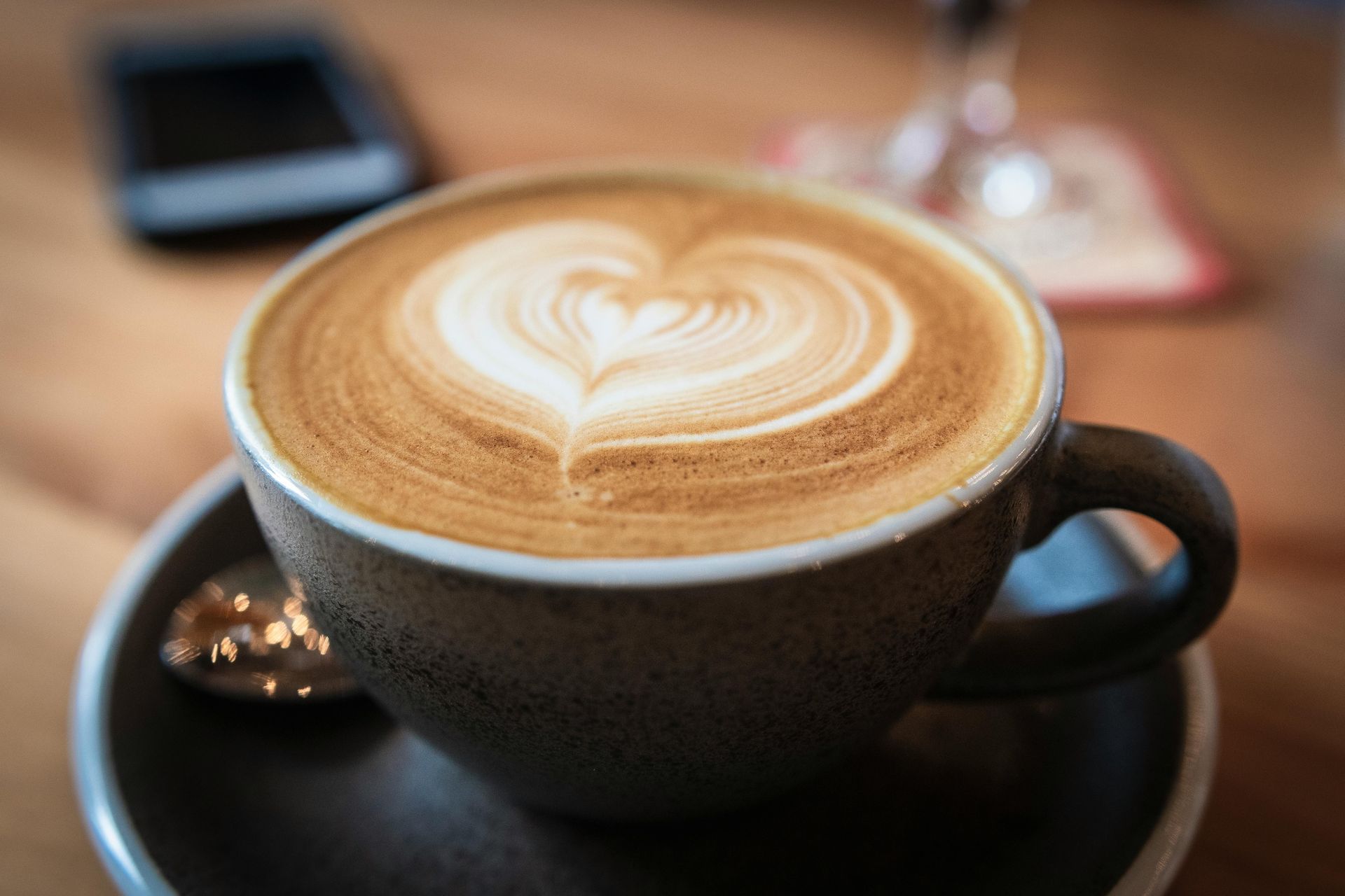 A cup of latte with heart-shaped foam art on a saucer. A smartphone and coaster are in the blurry background.