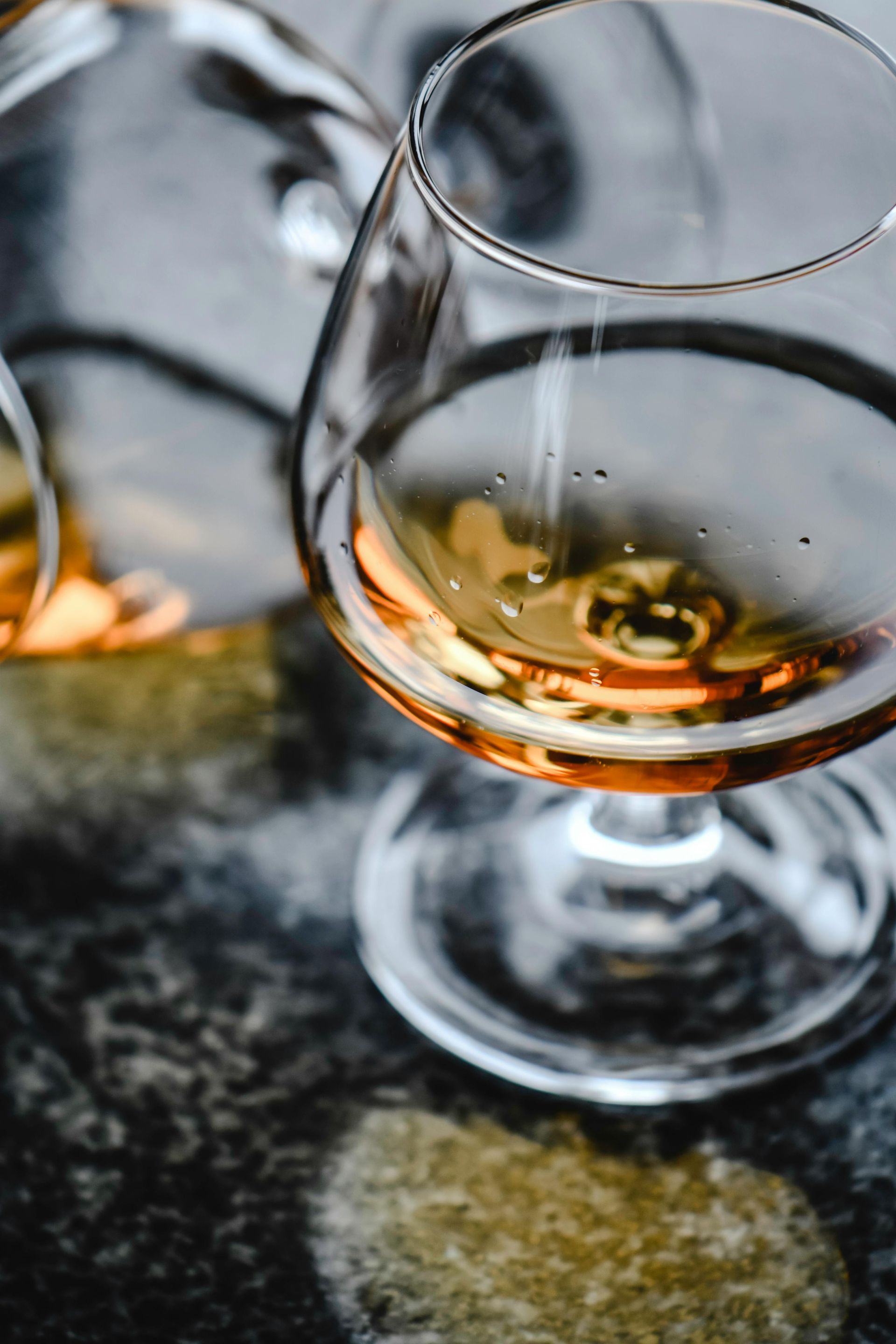 Two brandy glasses filled with amber liquid on a mottled gray surface, one in sharp focus.