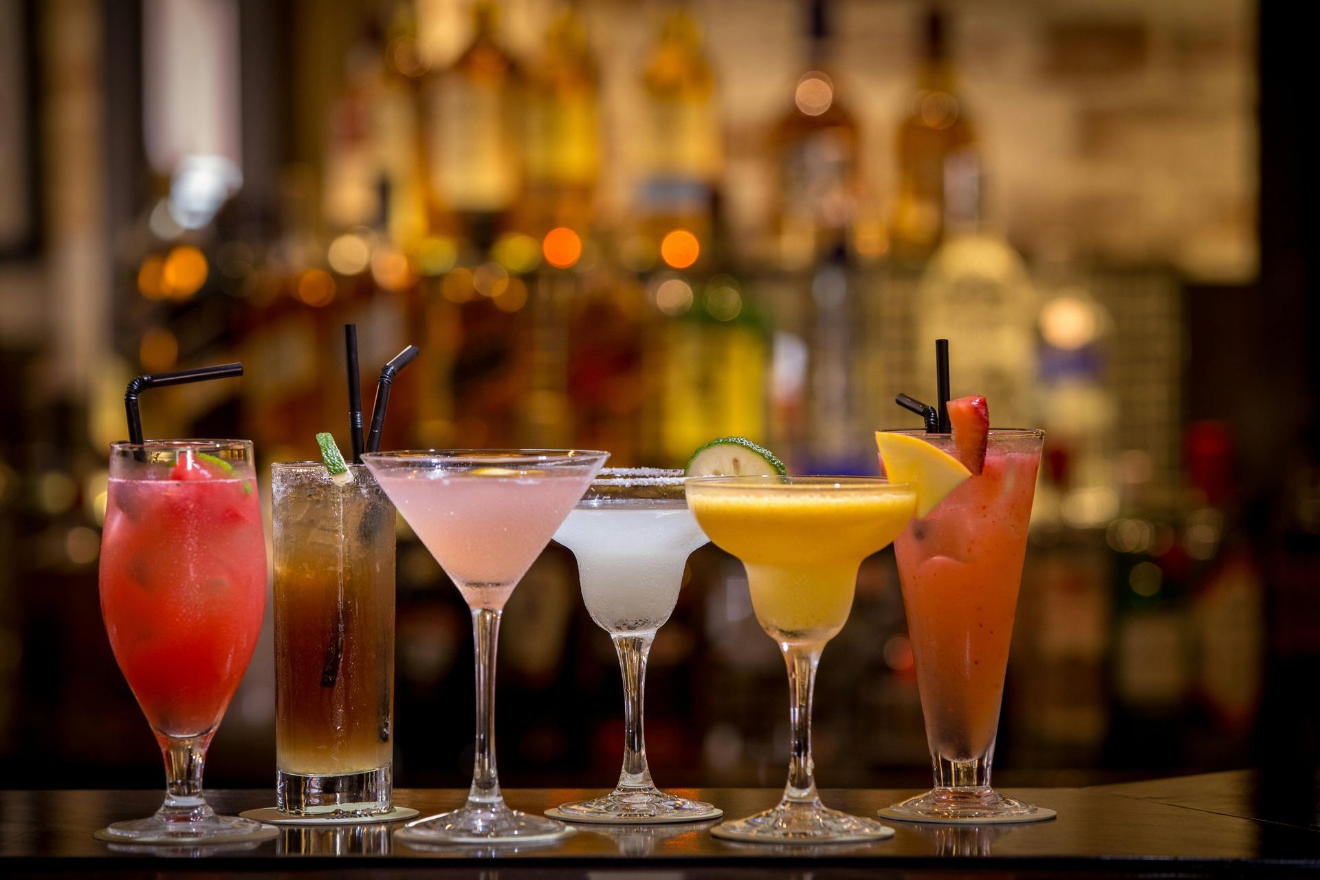Six colorful cocktails on a bar, with liquor bottles blurred in the background.
