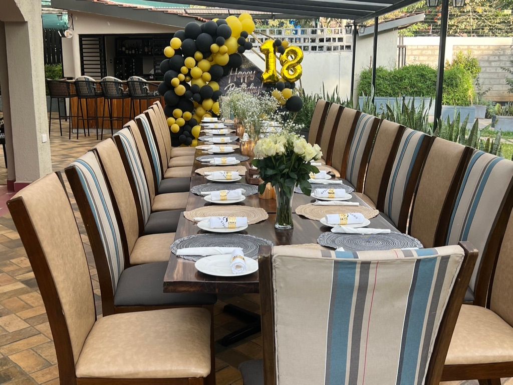 Long table set for a party, decorated with balloons, flowers, and place settings. Outdoors, chairs with patterned backs.