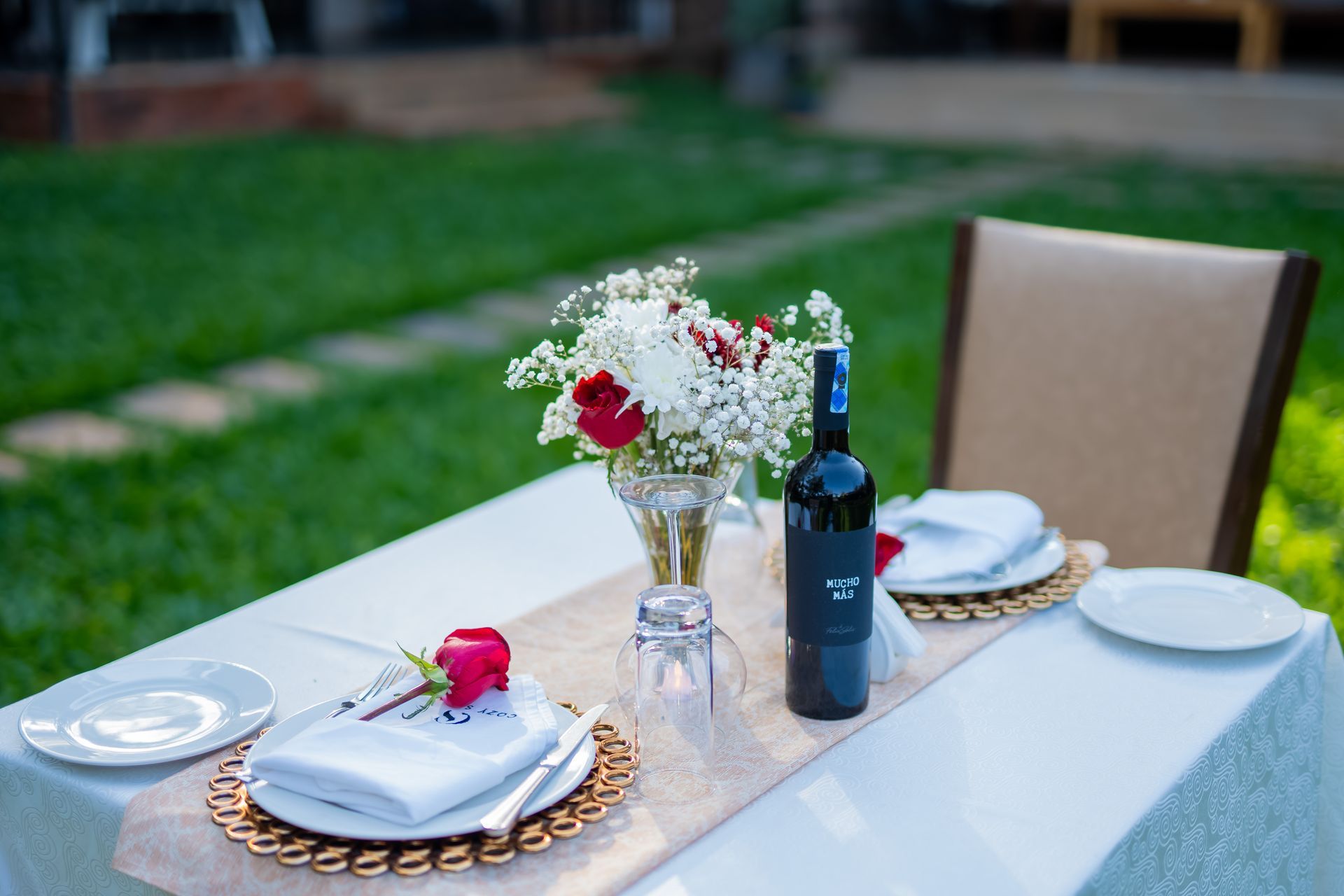 A table setting outdoors with a floral centerpiece, wine and place settings on a white tablecloth. Green grass in background.