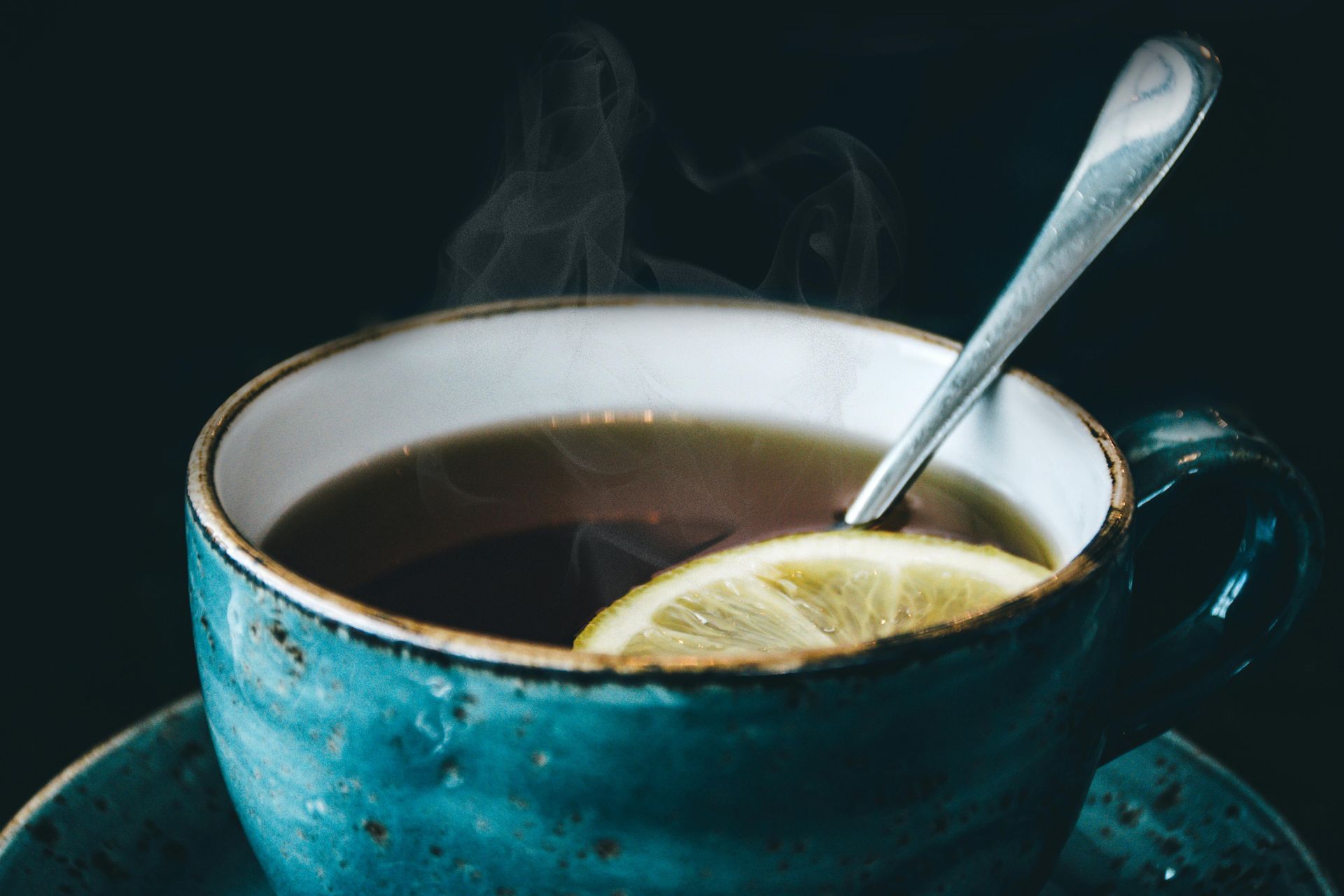 Steaming cup of tea with a lemon slice and spoon, in a blue ceramic mug on a dark background.