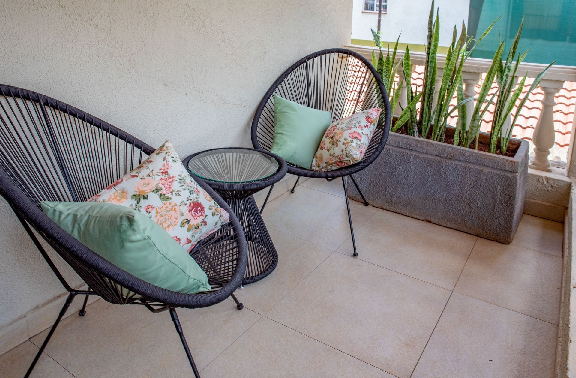 Two black wicker chairs with pillows and a small table on a balcony with a planter box of plants.