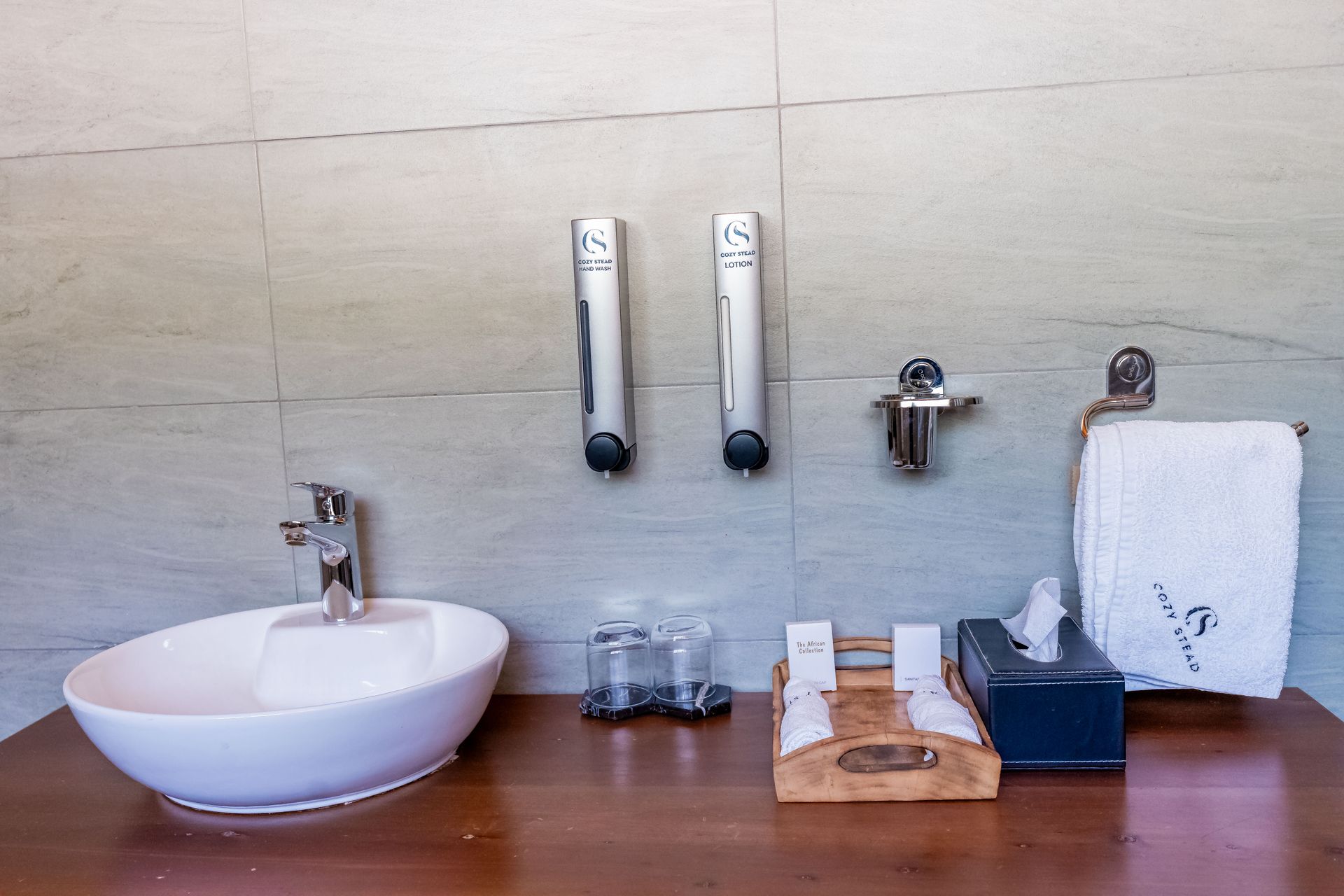 Bathroom sink area with a white sink, dispensers, and accessories on a wooden counter against a light-colored tiled wall.