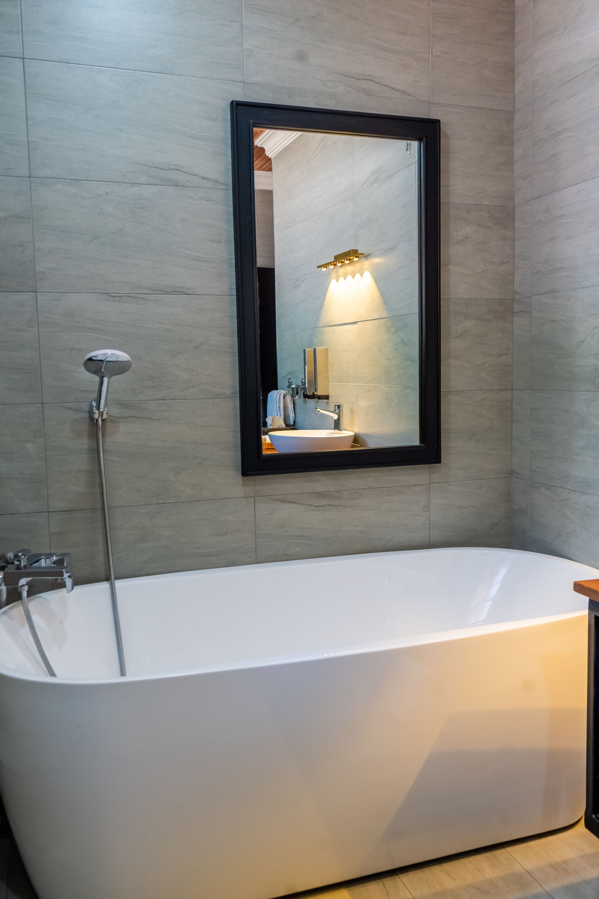 A white freestanding bathtub is below a black-framed mirror in a modern bathroom with gray tiled walls.