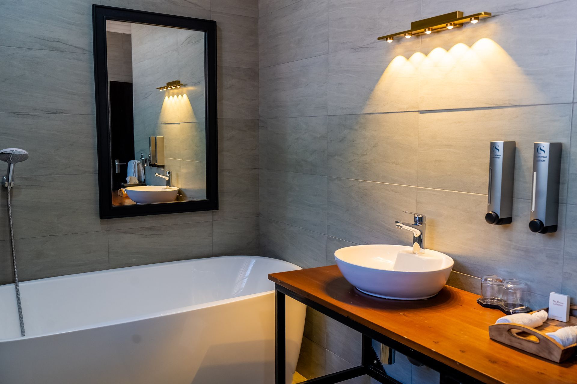 Bathroom with a white tub, a wooden countertop with a sink, and a mirror reflecting the scene. The walls are gray tile.