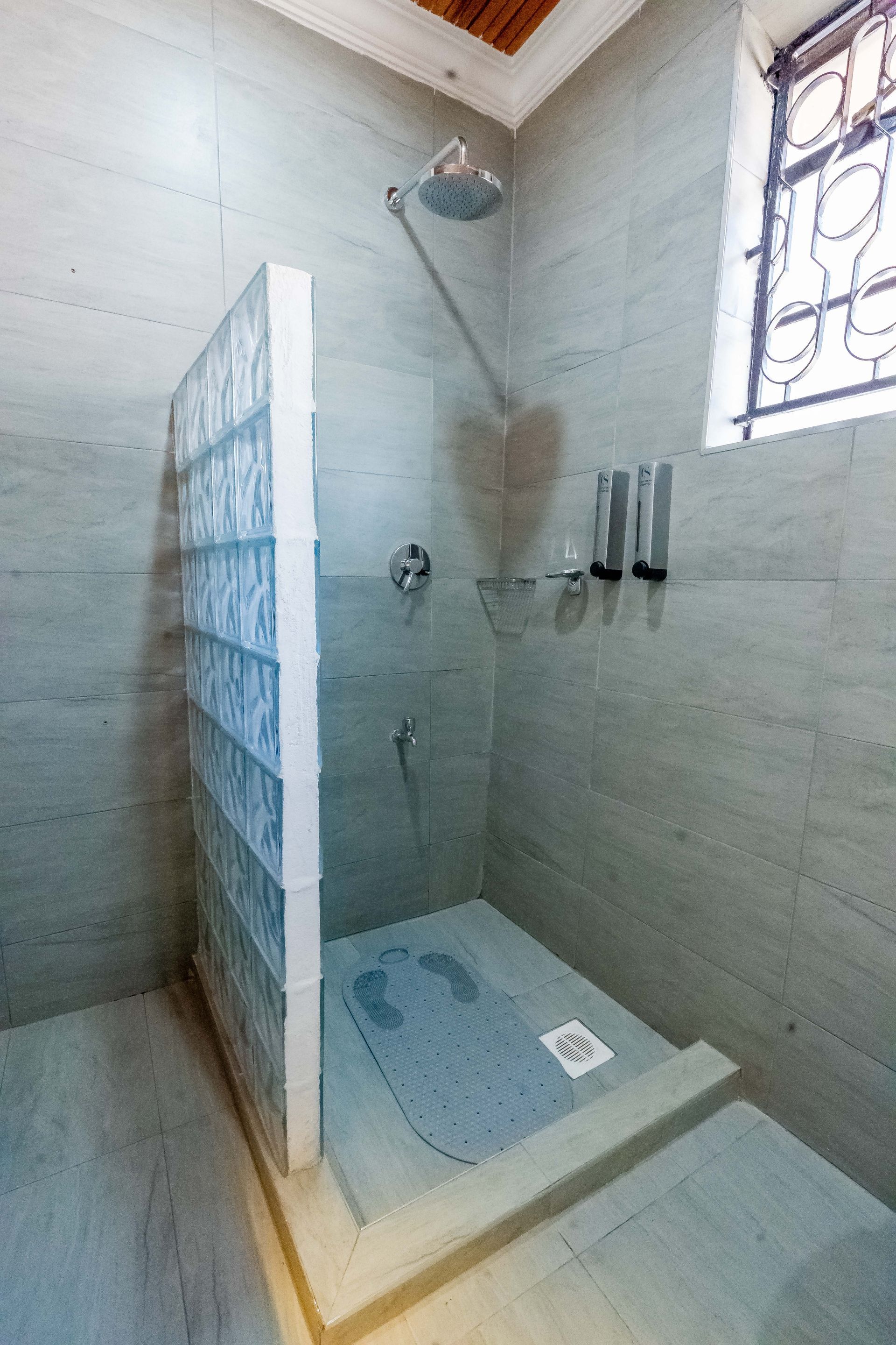 Shower stall with glass block wall, gray tile, rain shower head, and soap bottles. A window is on the right.