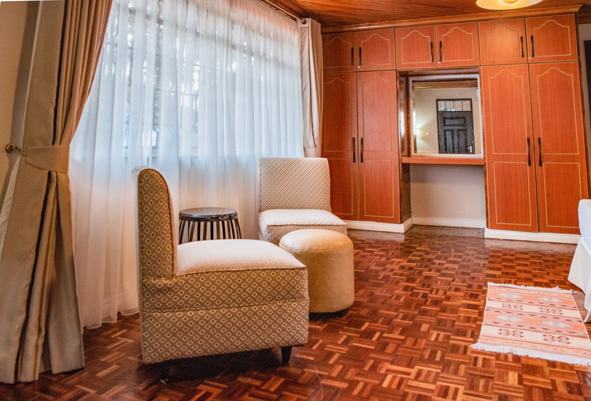 A cozy bedroom with parquet flooring, wooden cabinets, a window with curtains, and two patterned chairs.