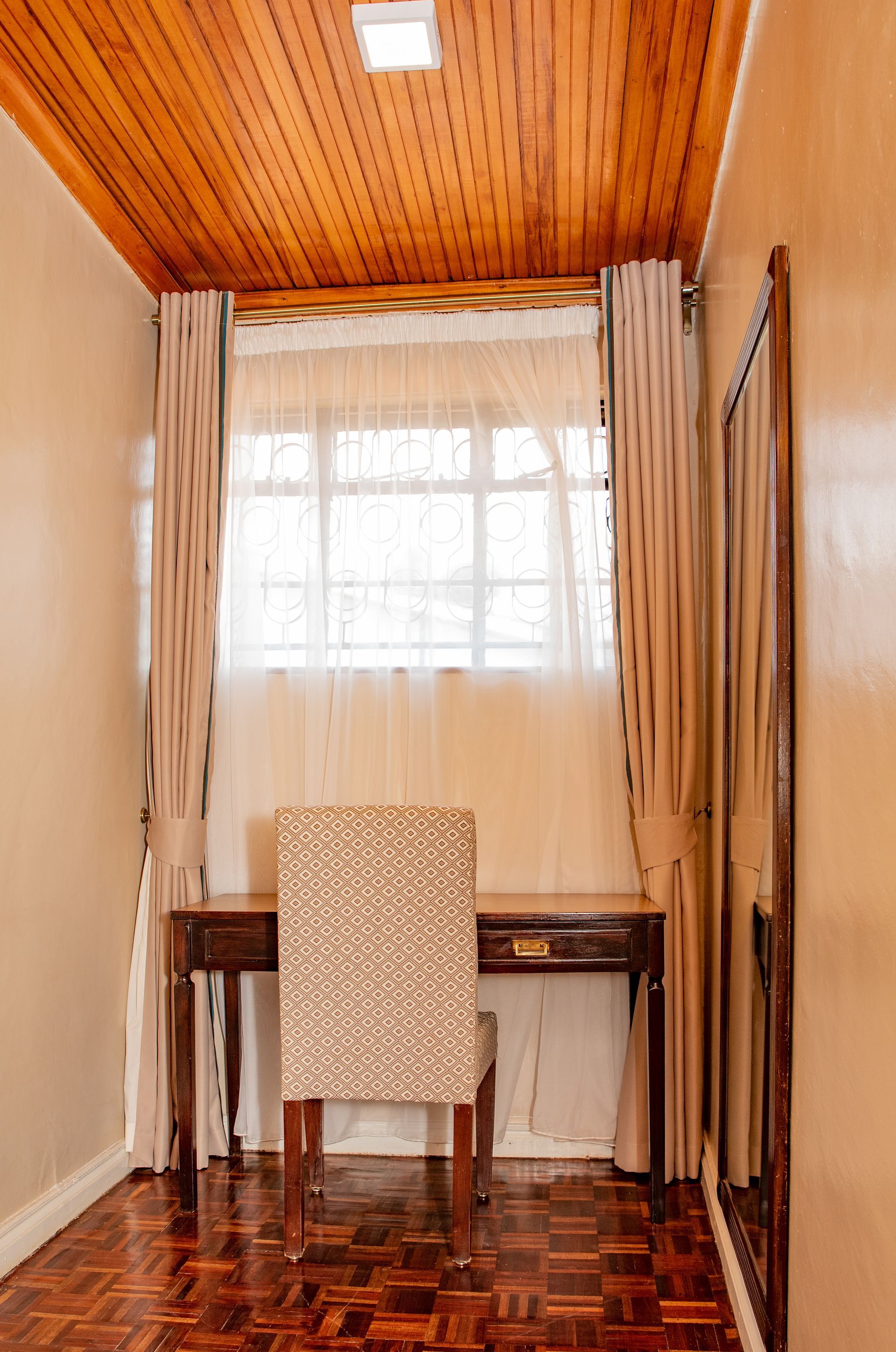 A office nook featuring a desk, patterned chair and window with curtains. The room has a wood ceiling and floor with walls.