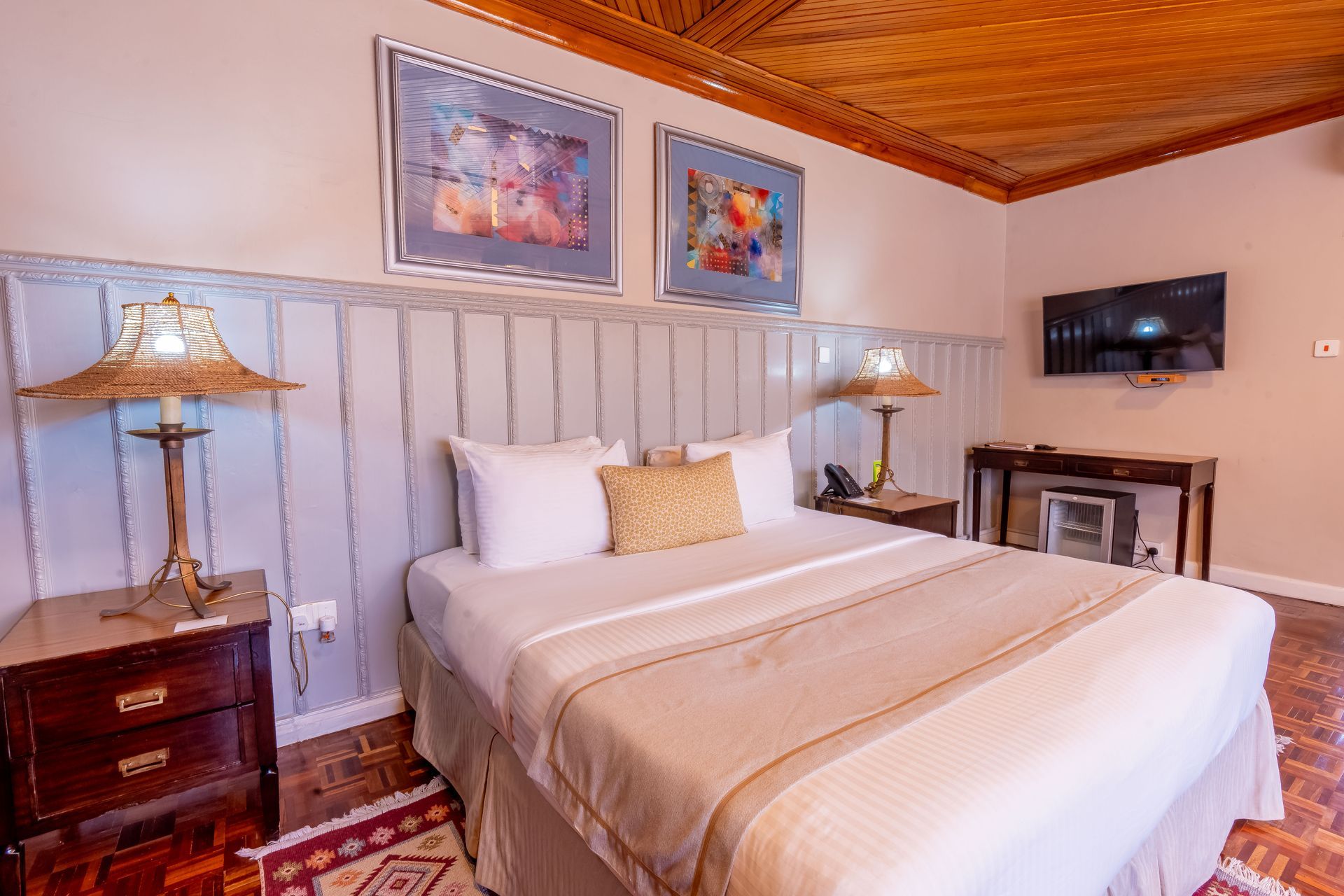 Bedroom with a bed, lamps, and a wall-mounted TV. Artwork hangs above the bed. The ceiling has a decorative wooden pattern.