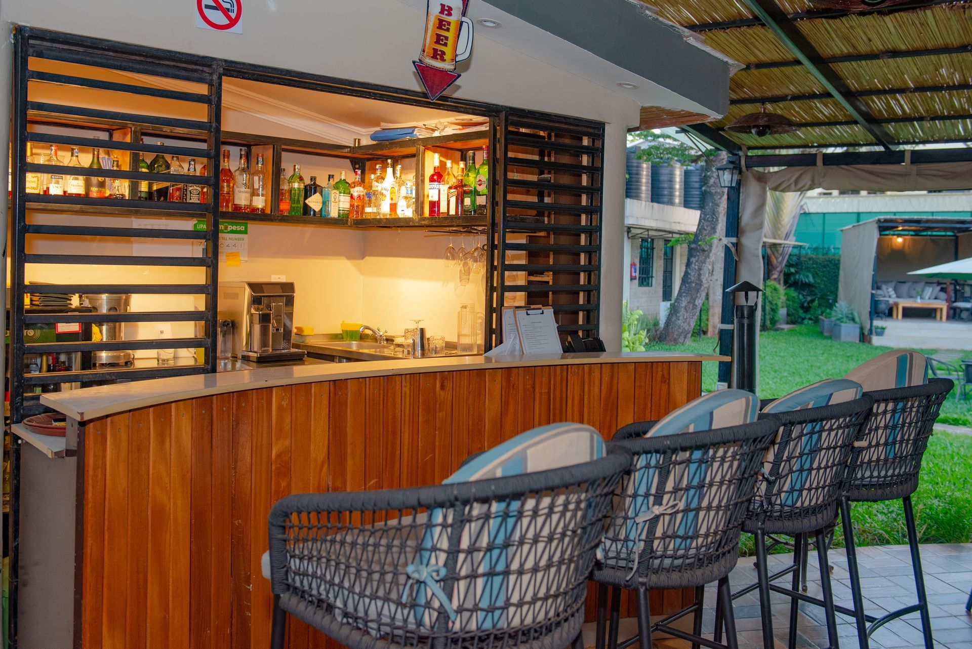 Outdoor bar with wooden counter and bar stools. Shelves hold bottles of liquor, and a coffee machine is visible.
