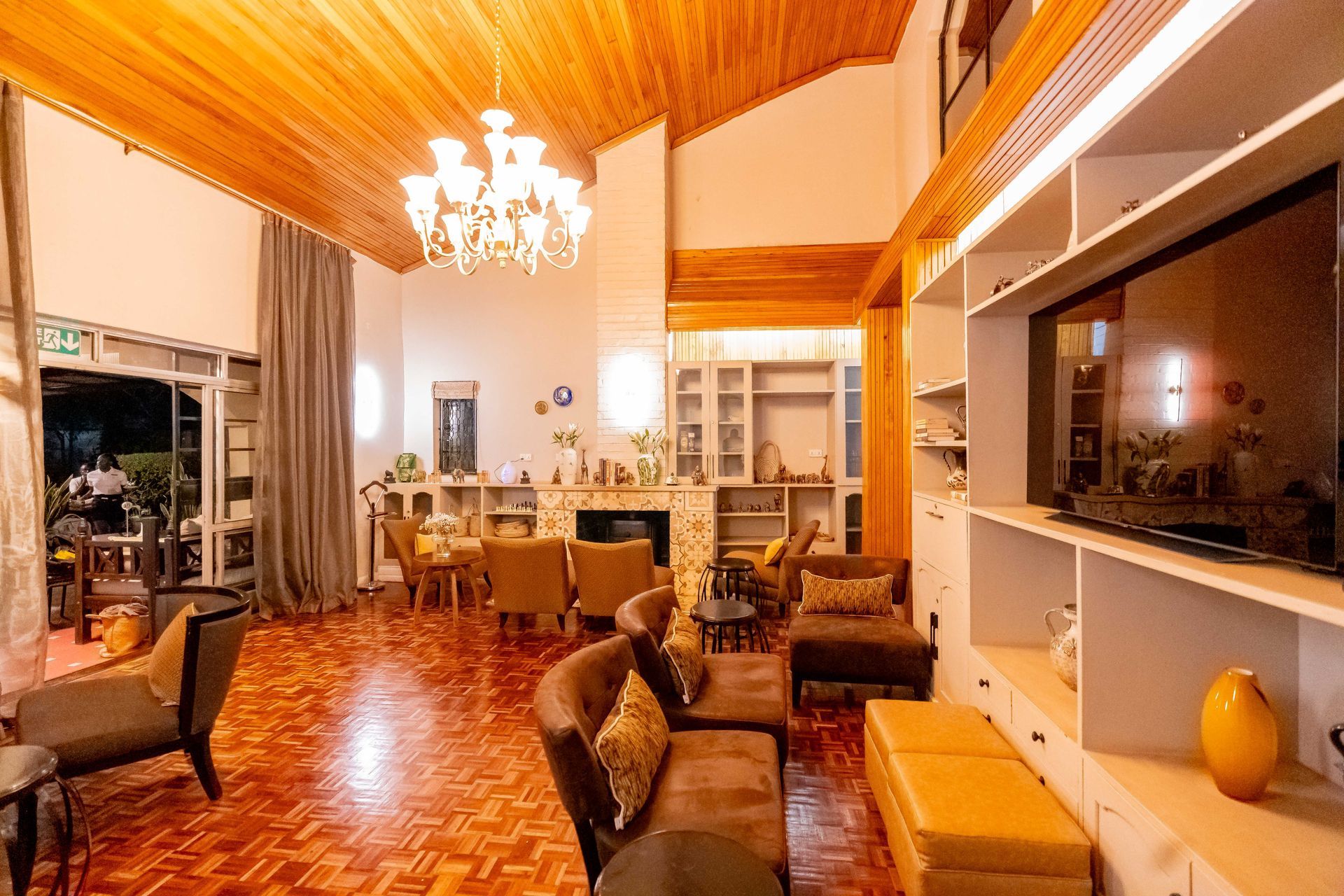 a lounge  with wood floors and ceiling. Furniture includes sofas and a TV and a chandelier. There is a fireplace and shelves.