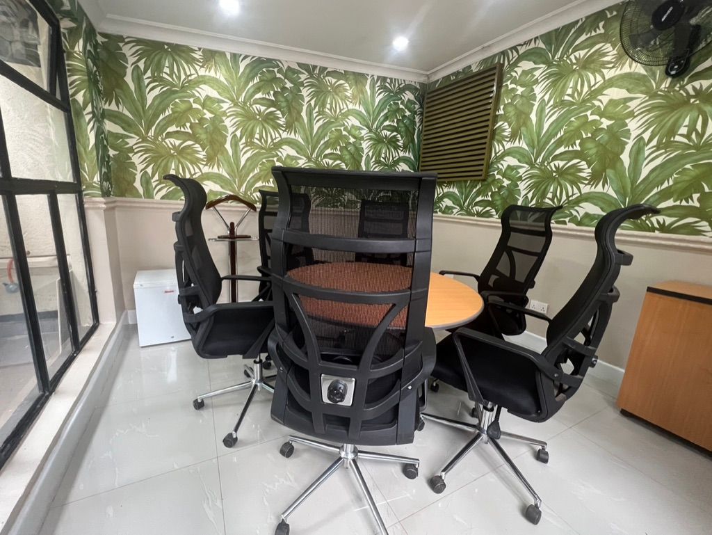 Office space with round table and black mesh chairs. Fern wallpaper, mini-fridge, and wood-toned cabinet.
