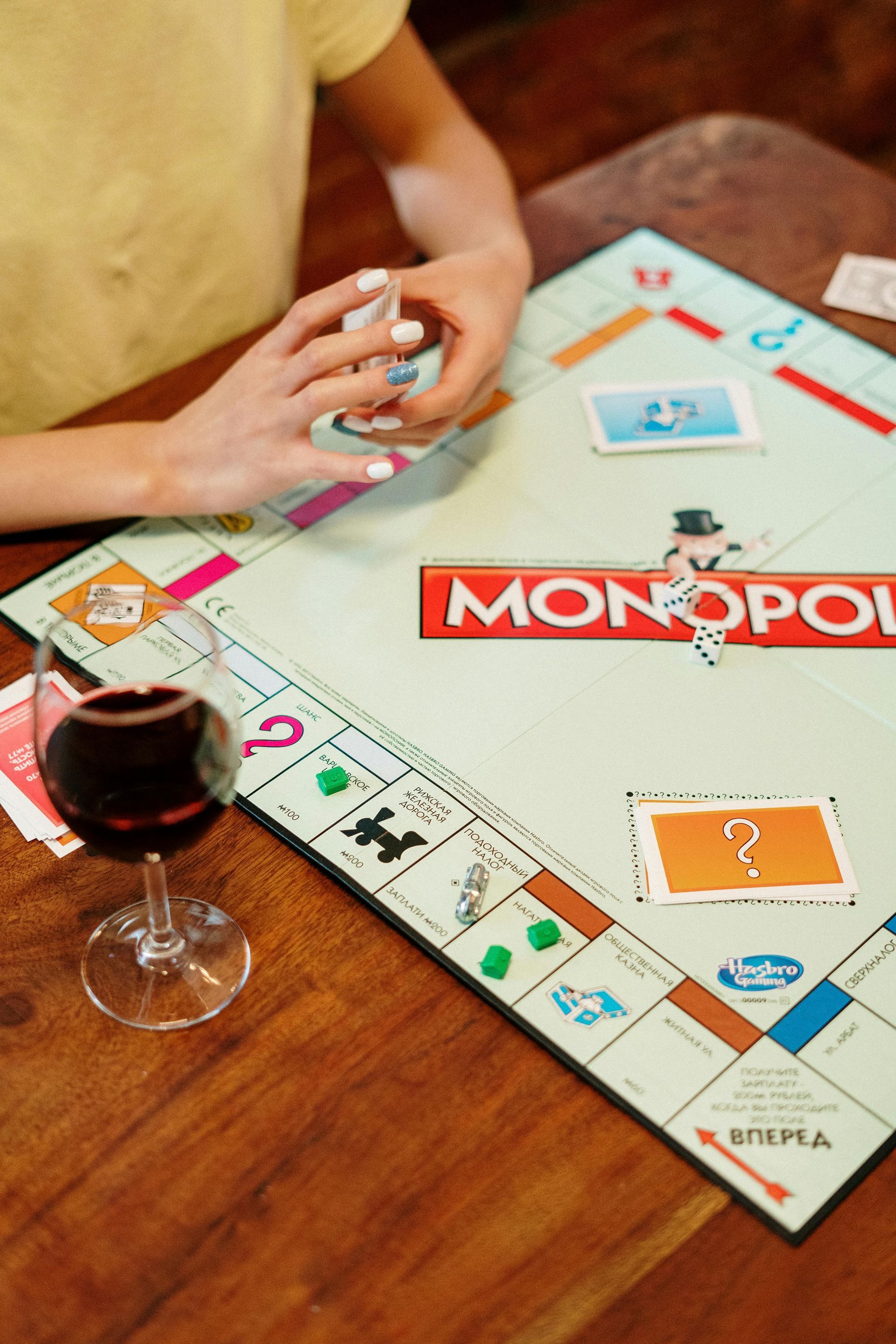 Person playing Monopoly, holding cards, with a glass of red wine on a wooden table.