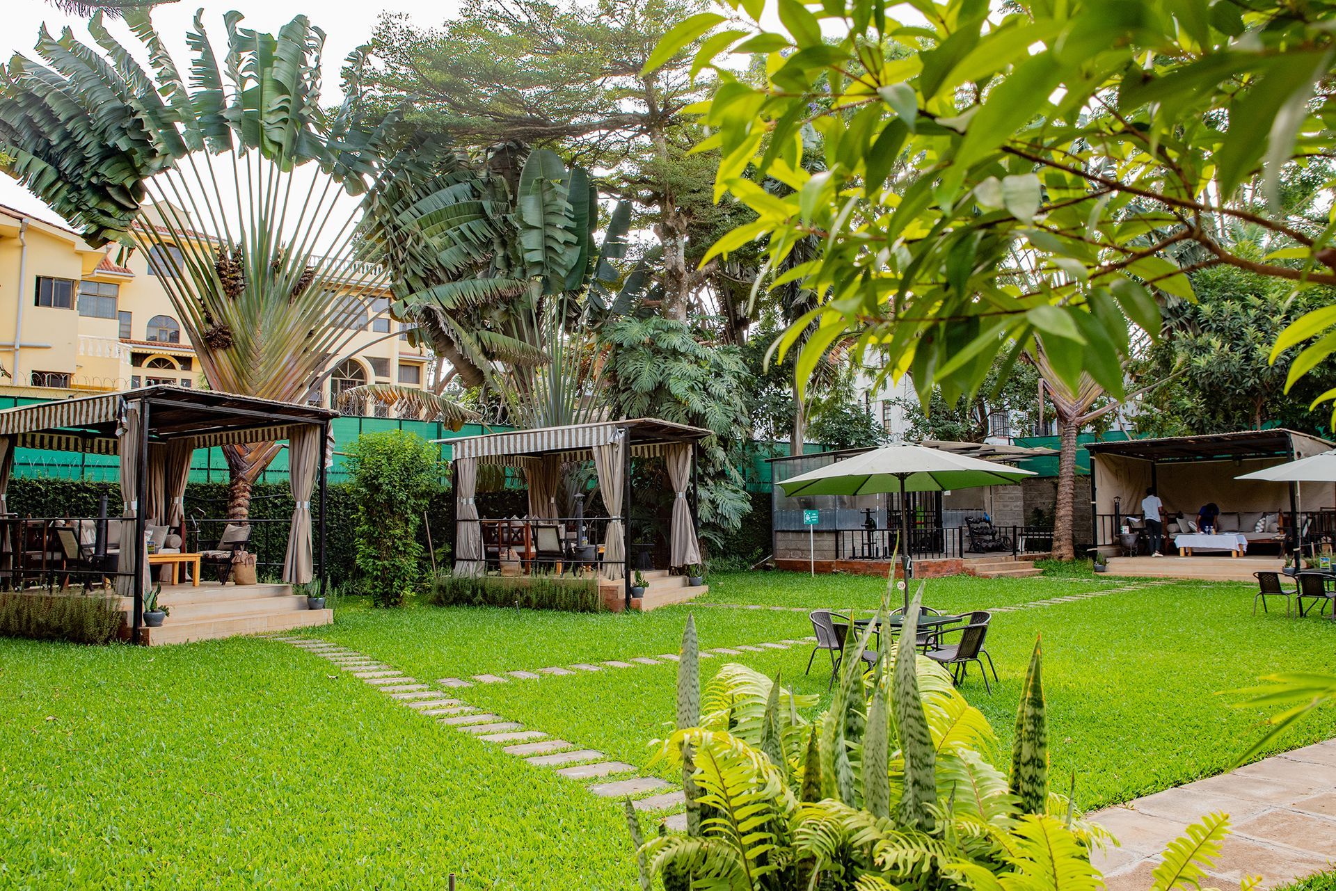 Lush green garden with gazebos, tables with umbrellas, and people dining. Buildings and tropical plants in the background.