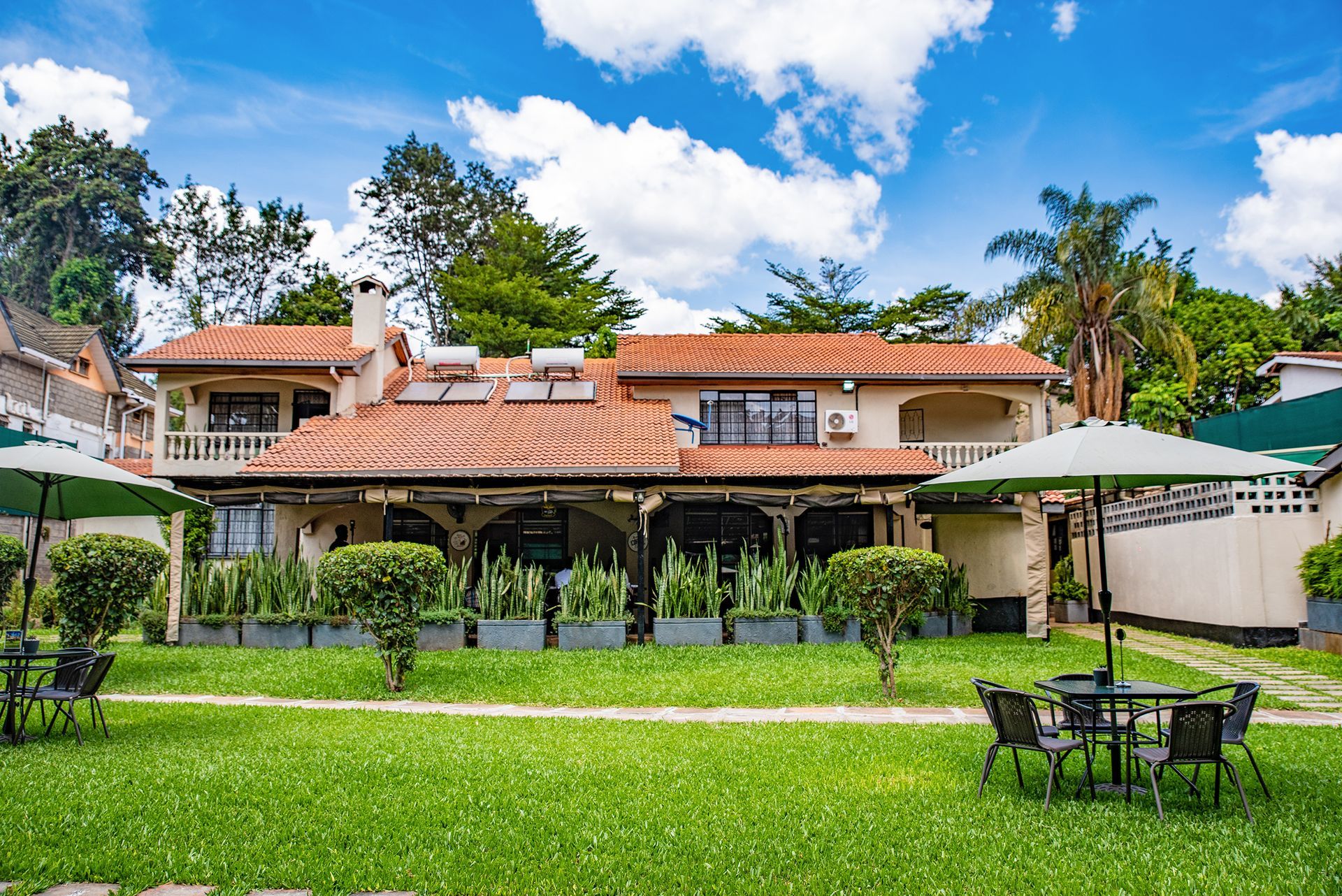 A two-story building with a terracotta roof, surrounded by a lawn with outdoor seating and shade umbrellas under a blue sky.