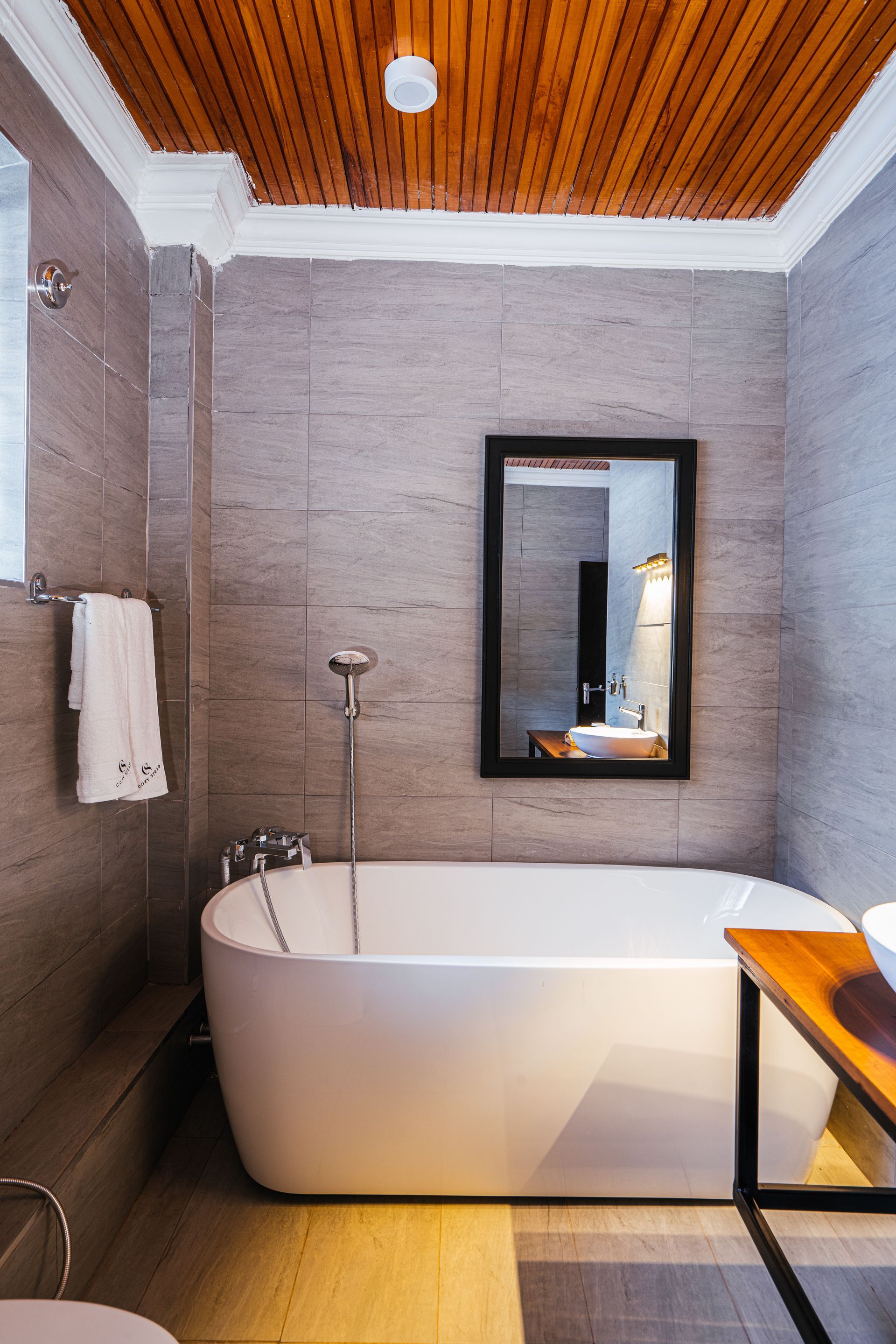 A bathroom with a white soaking tub, tiled walls and a wooden ceiling. A mirror hangs above the tub, and a towel on the left.