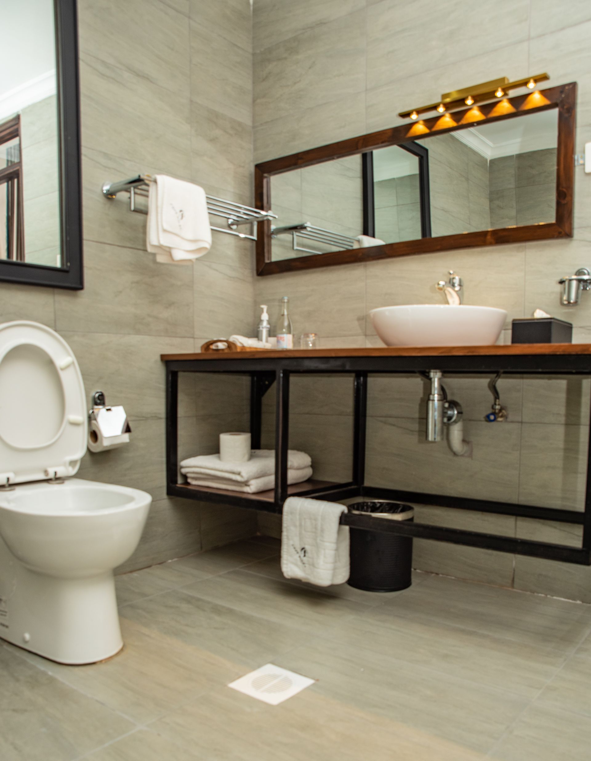 A modern bathroom with a toilet, sink, and mirror. Gray tile walls and a wooden vanity.