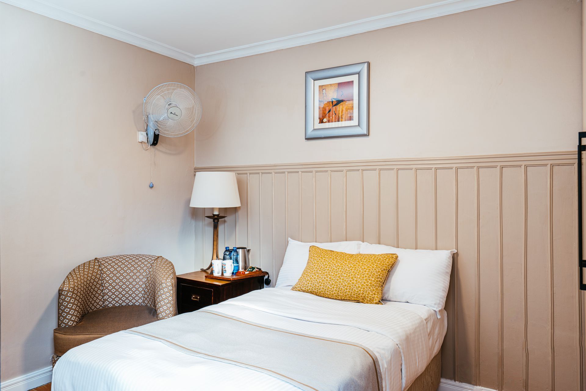 A small bedroom with a single bed, a side table, a wicker chair, and wall art; painted in beige and white.
