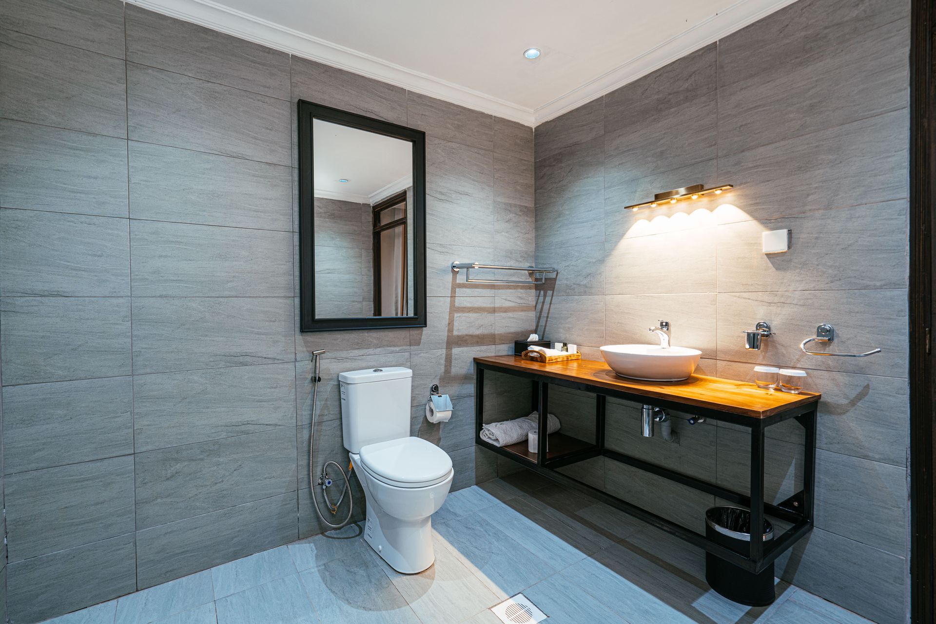 Modern bathroom with gray tiled walls, a toilet, sink on a wooden counter, and a large mirror.