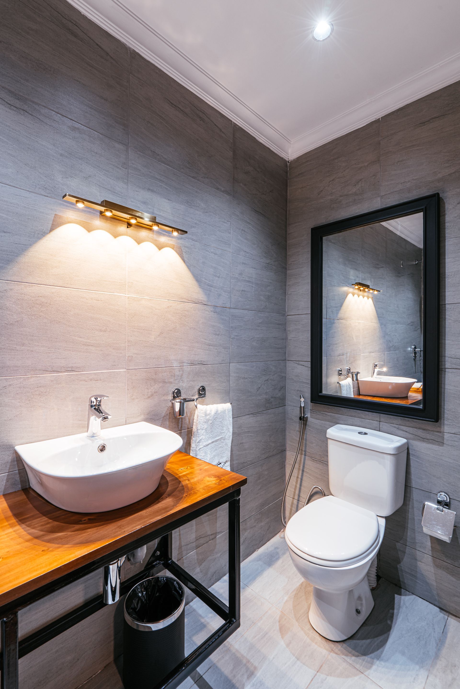 A small, modern bathroom with a white sink, toilet, and a black-framed mirror. Gray tiled walls and a wooden counter.
