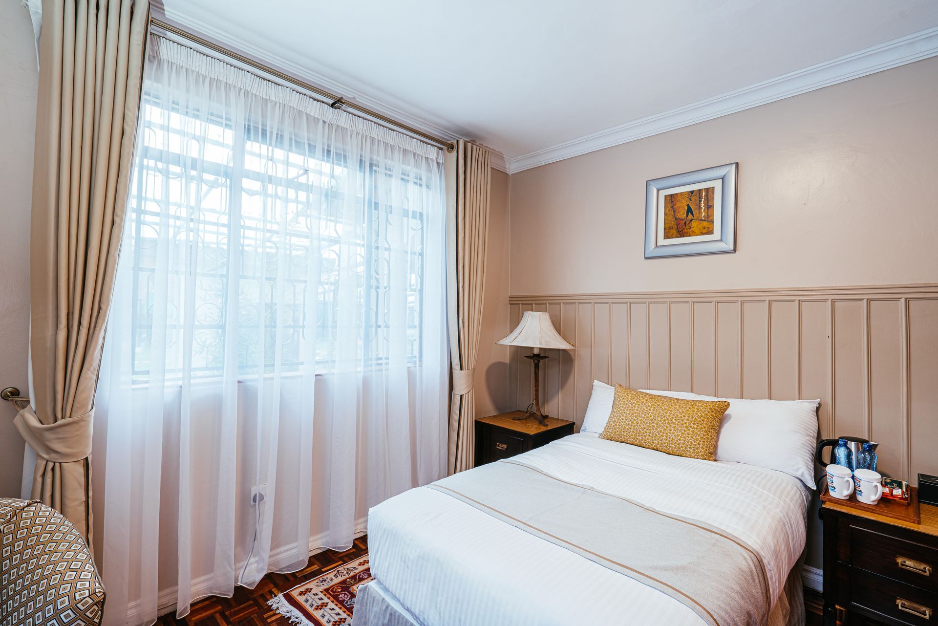 A bedroom with a bed, window with curtains, and a nightstand . The walls are beige and the bed linens are white and gold.
