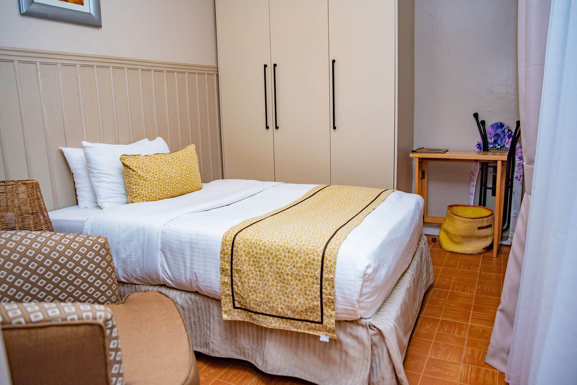 A bedroom with a bed, wardrobe, small table, and an armchair. Gold and white bedding and patterned accents give a warm feel.