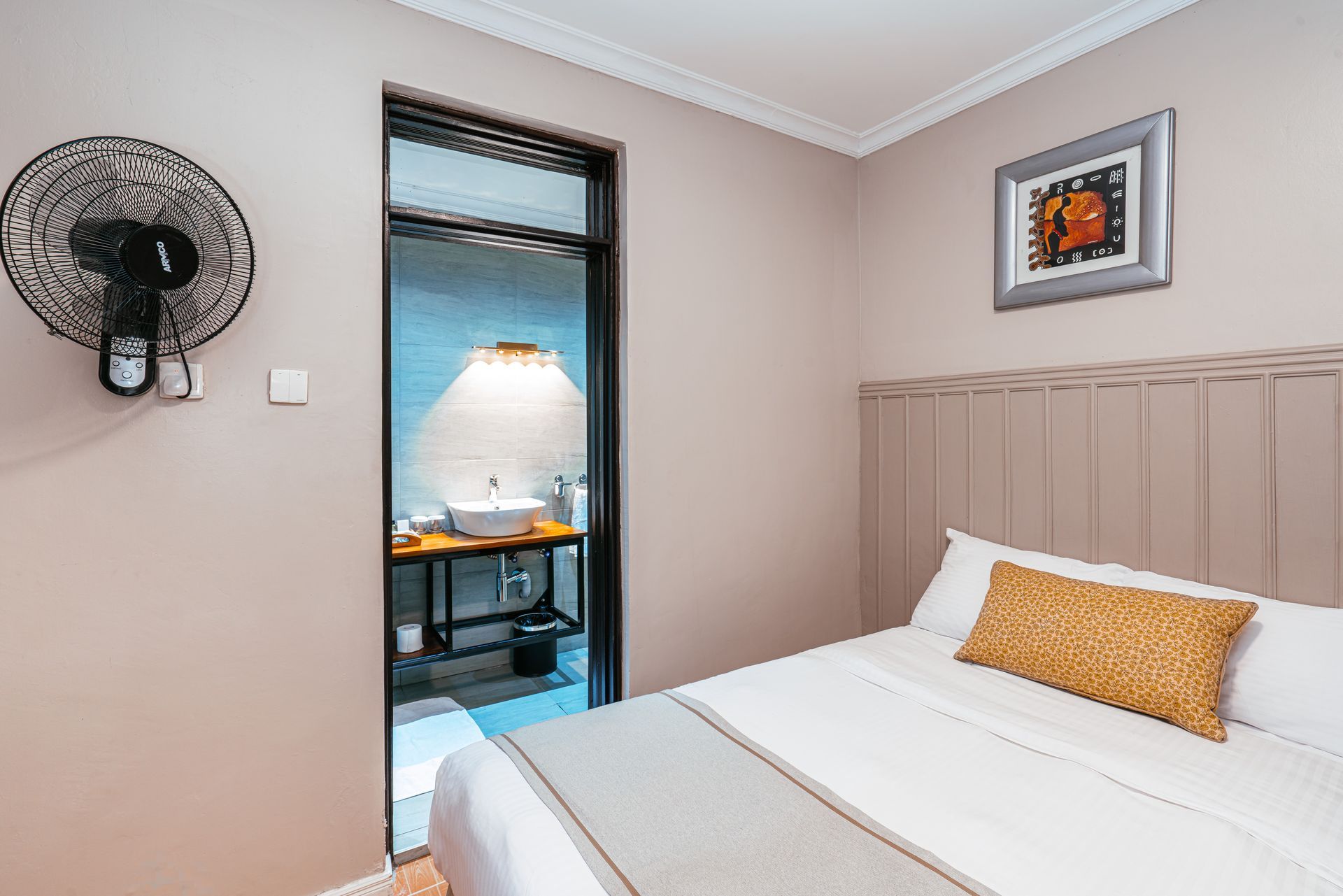 A bedroom with a bed, a bathroom entrance, and a wall-mounted fan. The walls are light pink, with a painting above the bed.