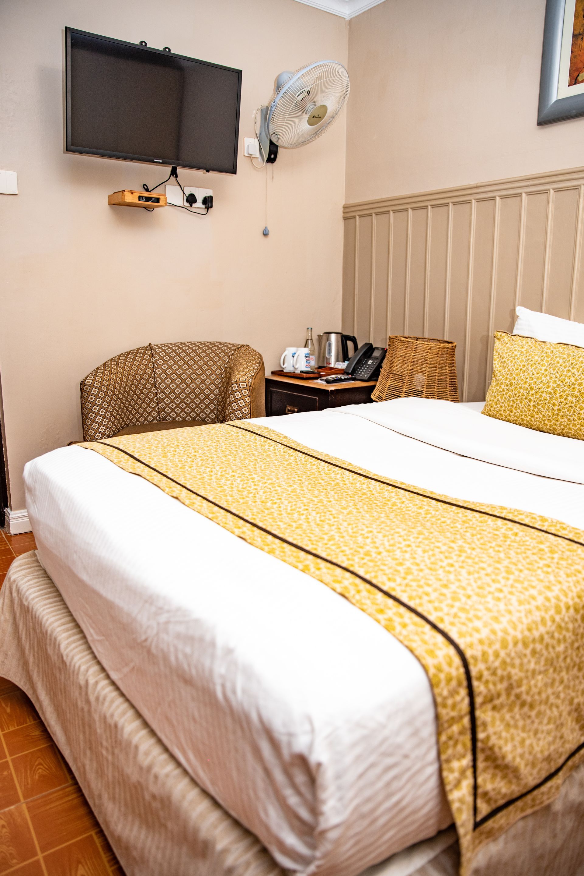 A hotel room with a bed, armchair, TV, and desk. The bed has yellow and white bedding, and the walls are beige.