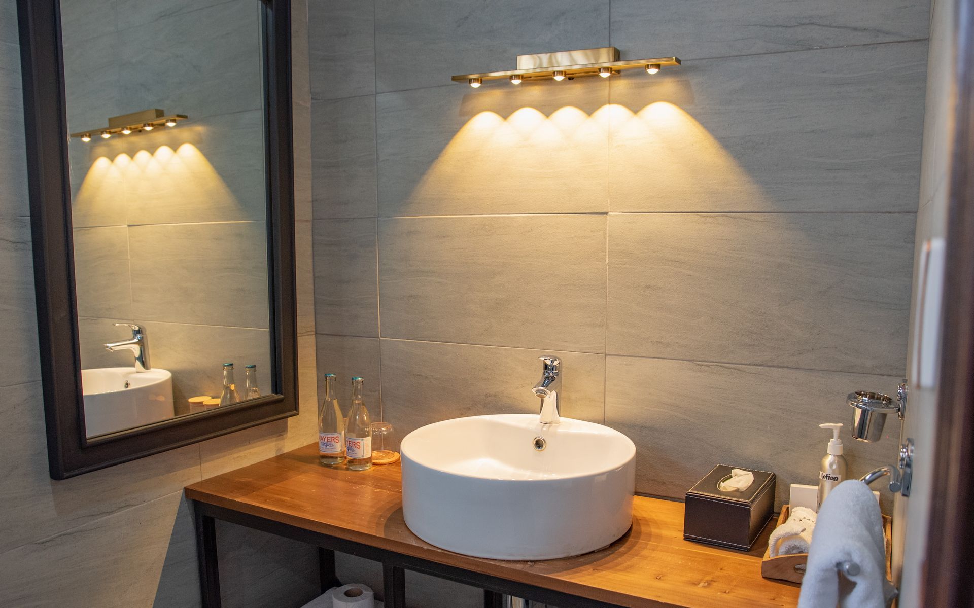 Bathroom with a white sink on a wooden counter, lit by a golden wall sconce. A large mirror reflects the sink and light.