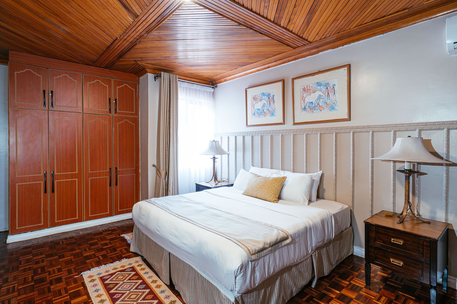 Bedroom with wooden ceiling, large wardrobe, bed with white bedding, two framed artworks, and bedside tables with lamps.