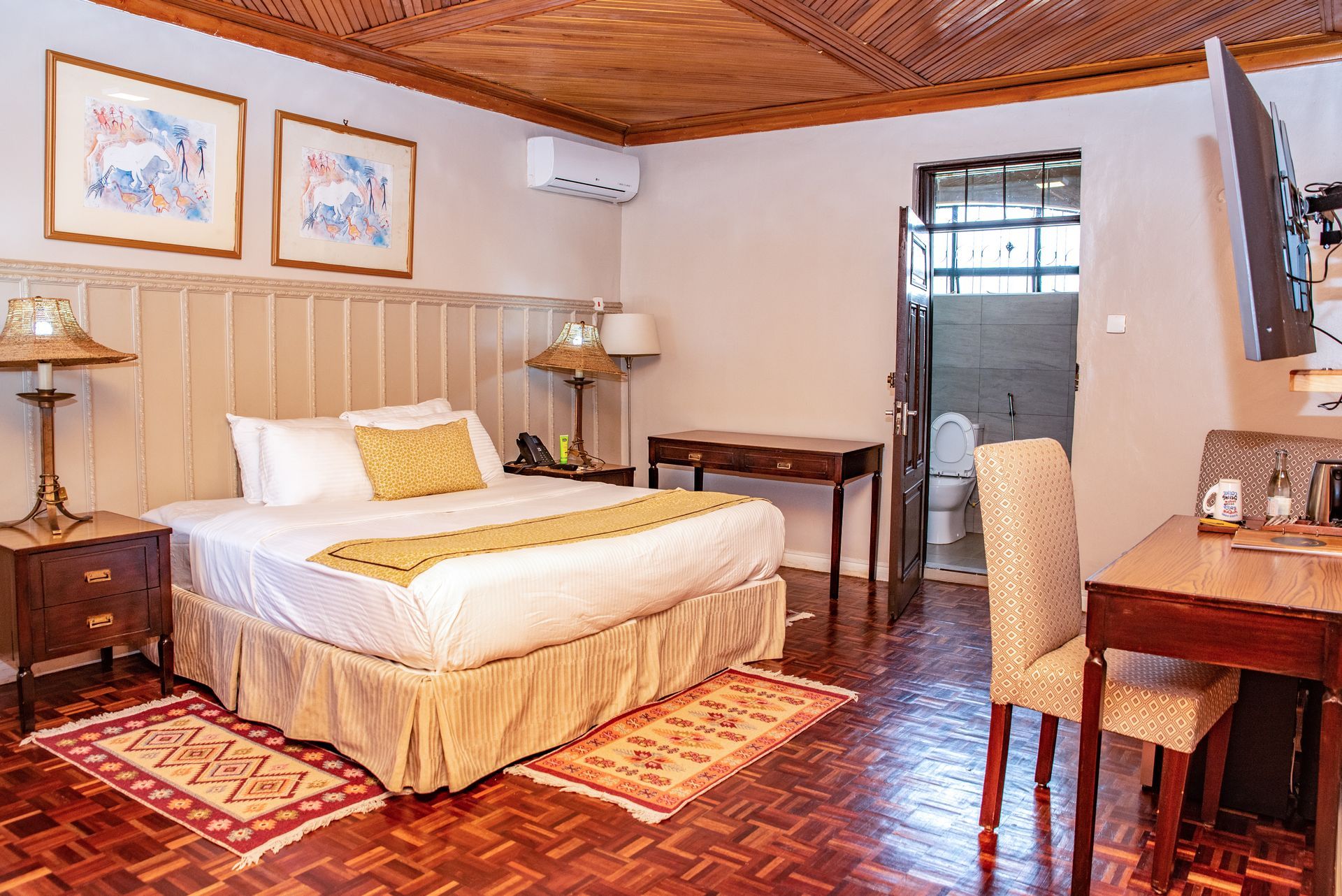 Hotel room with a bed, desk, and bathroom visible. Warm tones, wood ceiling, and framed art.