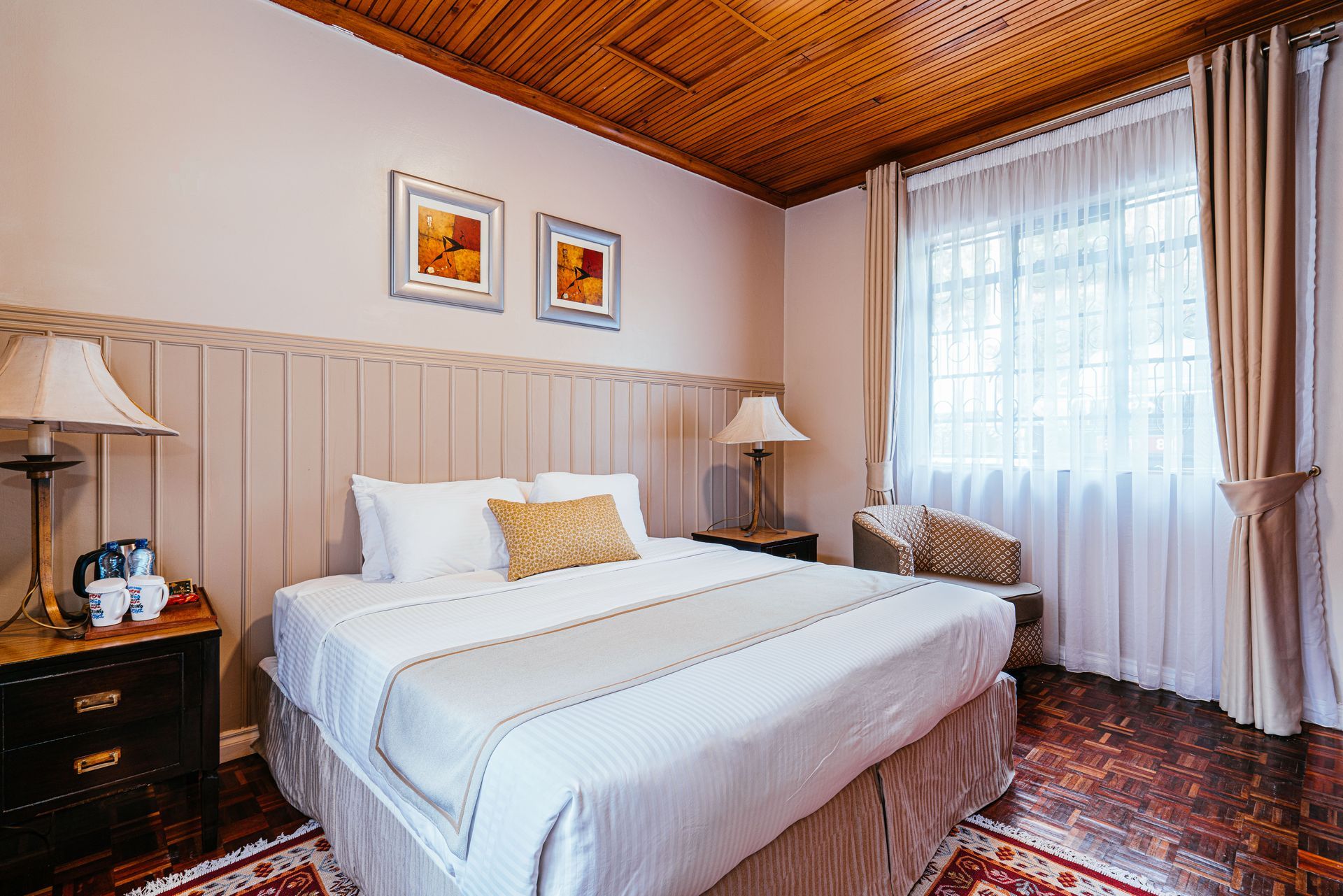 Bedroom with a bed, nightstands with lamps, ceiling, and window with curtains. The walls are beige, and the floor is dark.