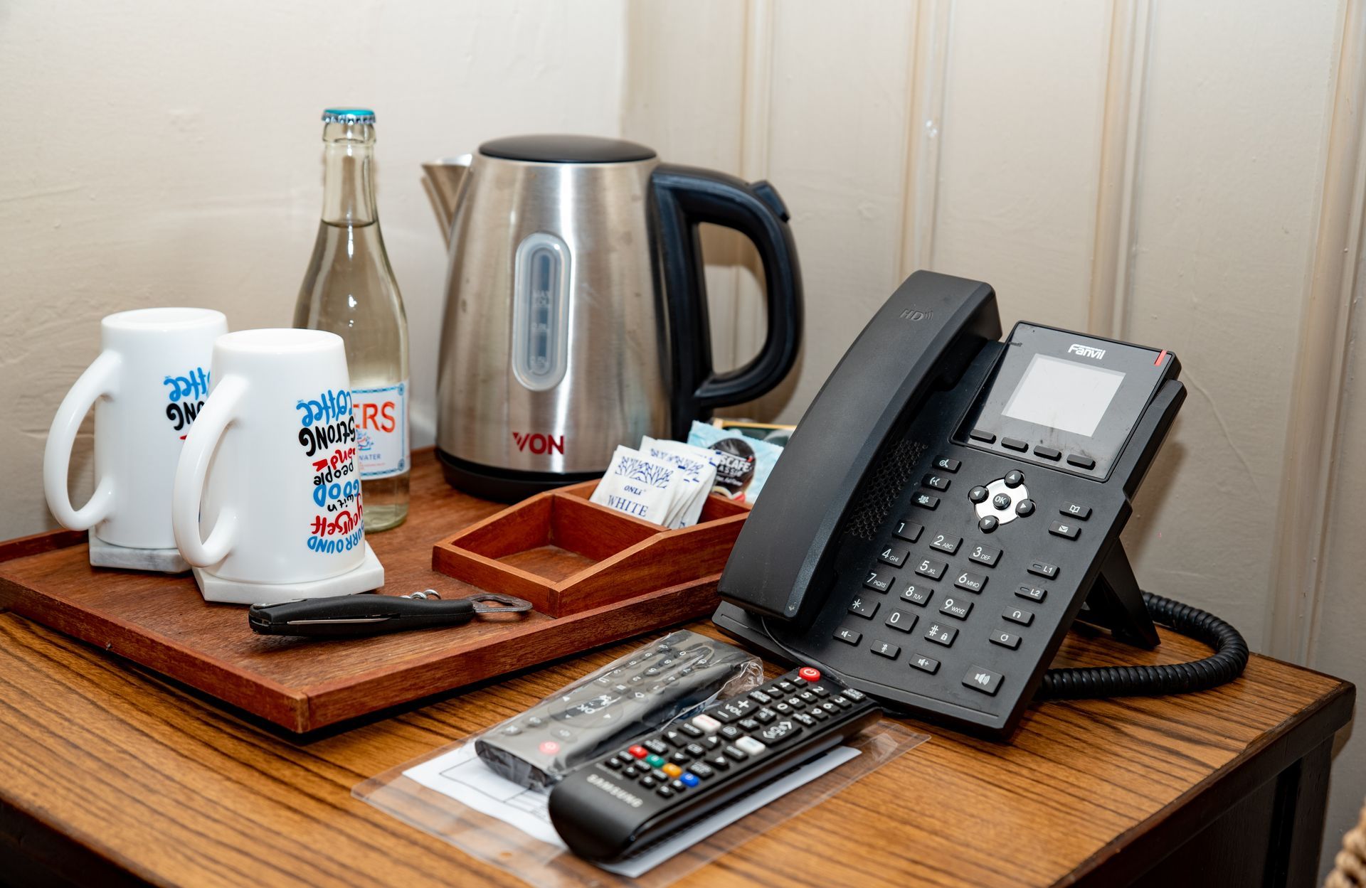A hotel room setup with mugs, a water kettle, a bottle, a phone, and remotes on a wooden surface.