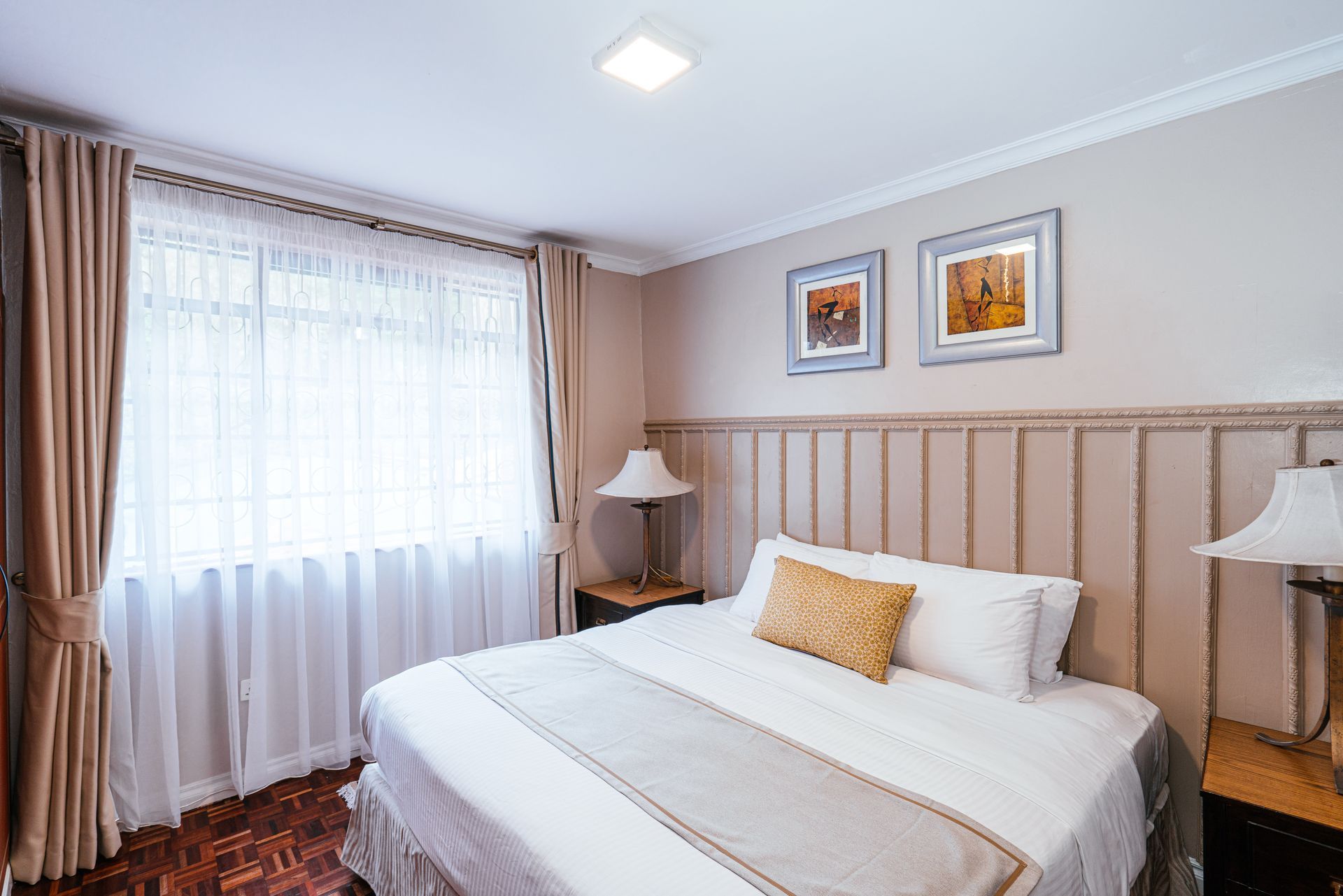 A bedroom with a bed, two lamps, a window with curtains, and framed art on the wall.