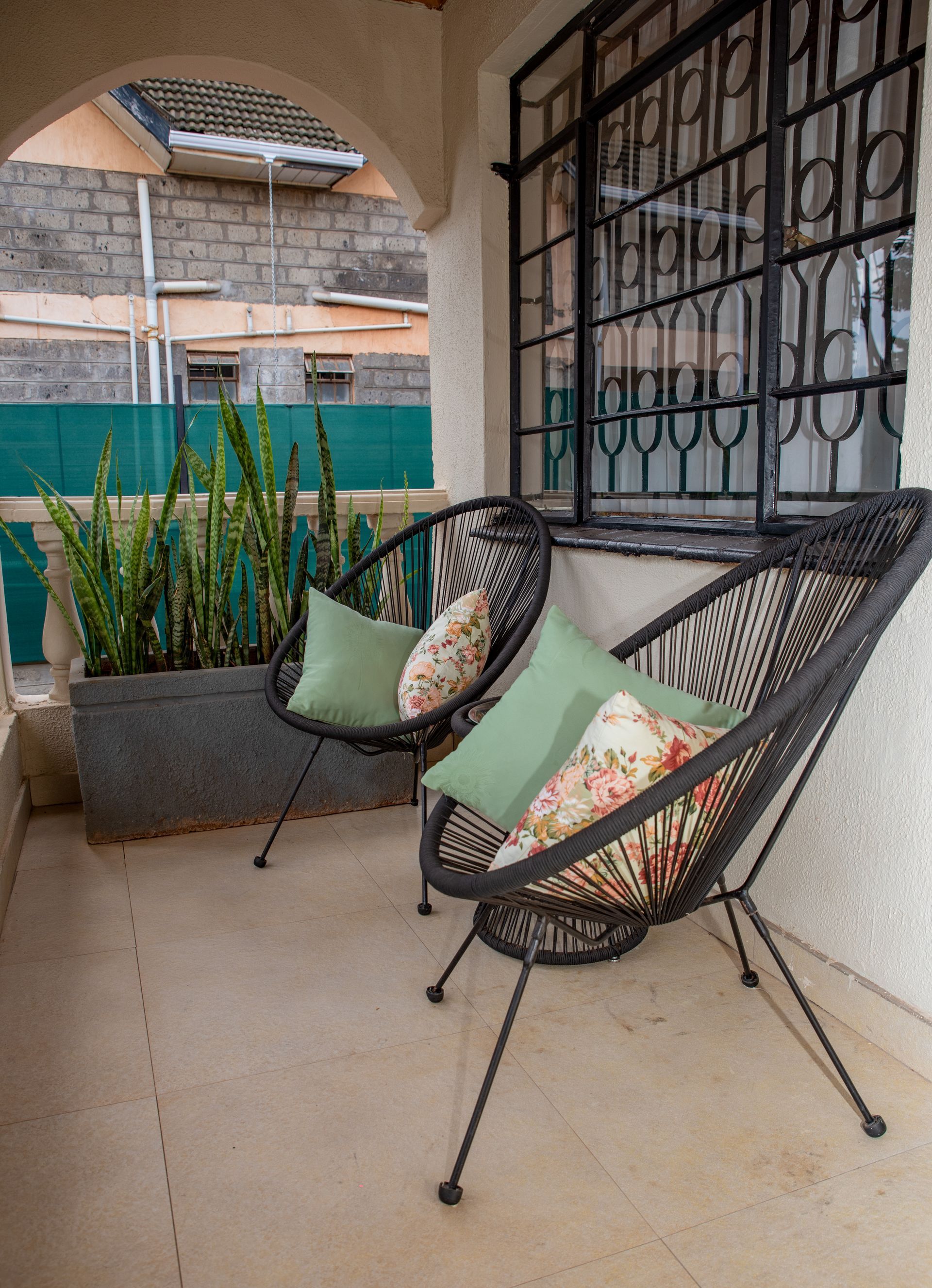 Two chairs with pillows sit on a porch next to a pot with tall plants. A window with decorative bars is behind the chairs.