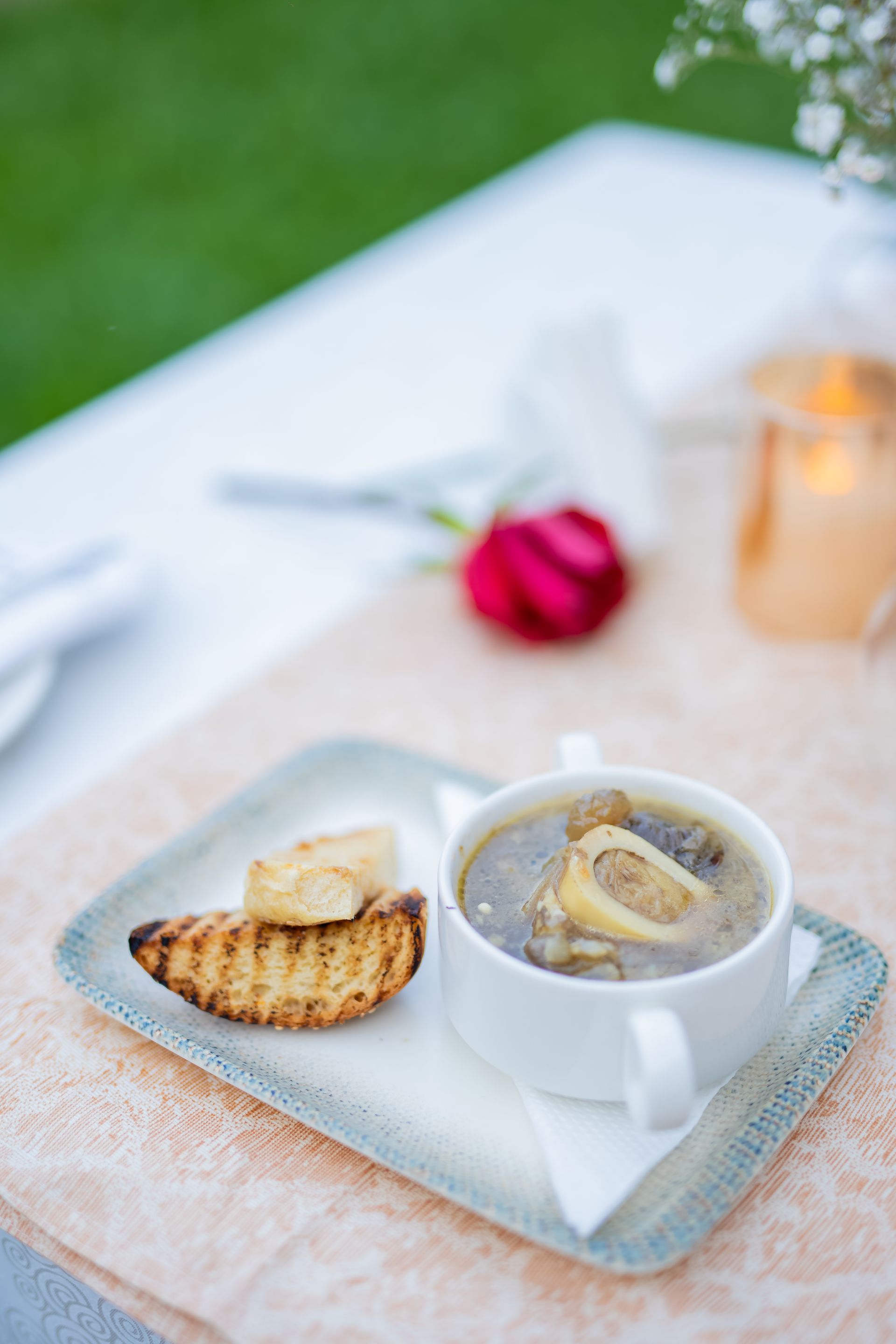 Bone marrow soup in a white bowl with toasted bread on a square plate, set on a white table outdoors with a rose.