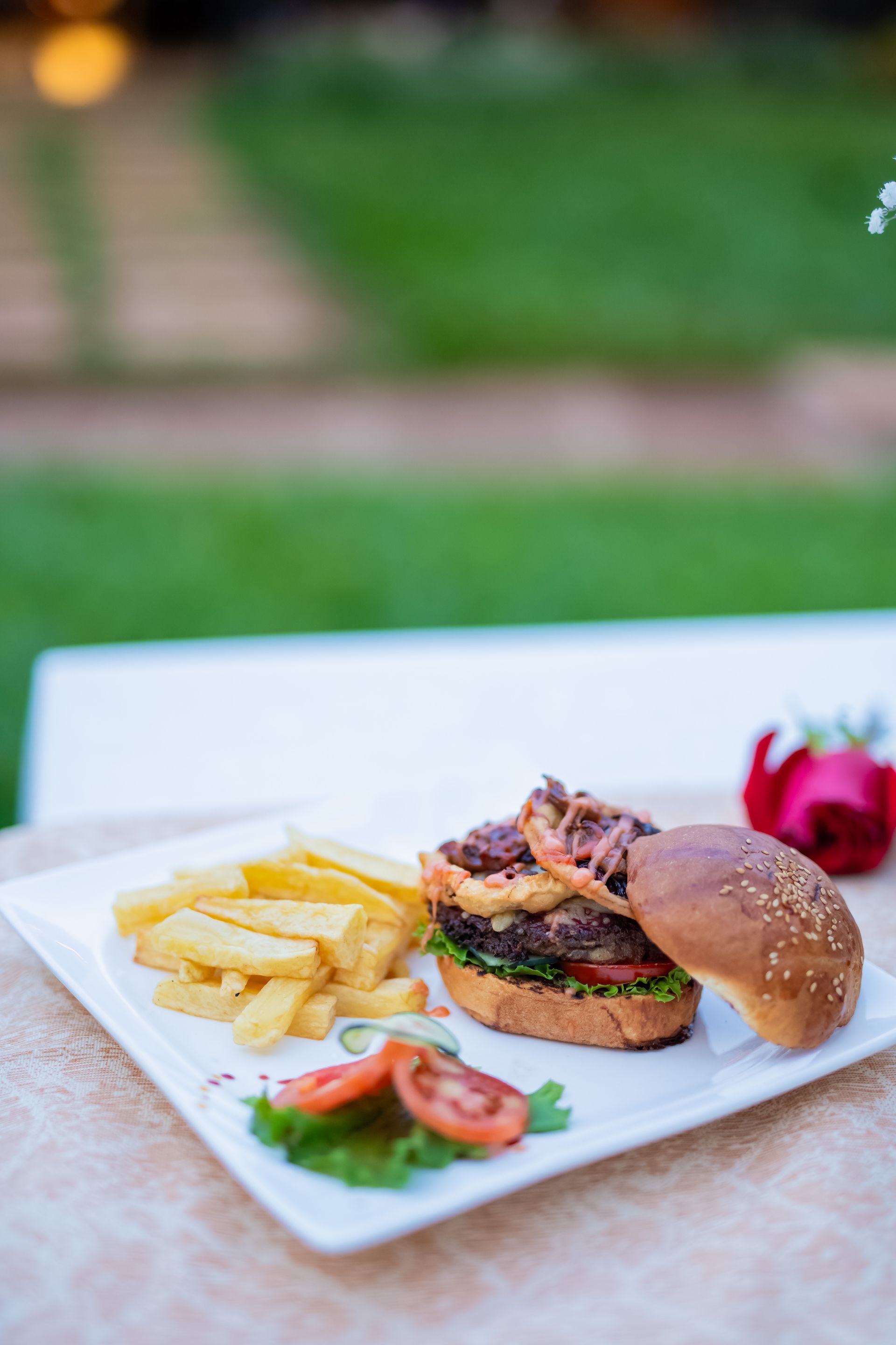 Hamburger with fries, tomato, and lettuce on a white plate outdoors. Burger has a sesame seed bun and onion rings.