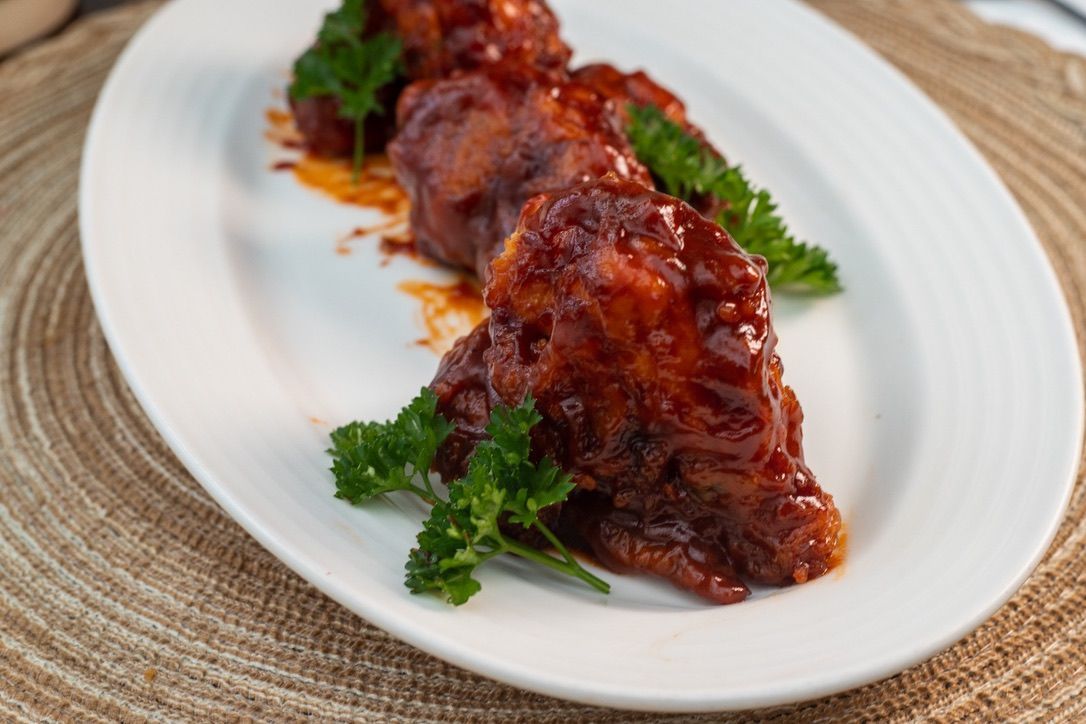Chicken wings coated in a red sauce are arranged on a white oval plate, garnished with parsley, atop a woven placemat.