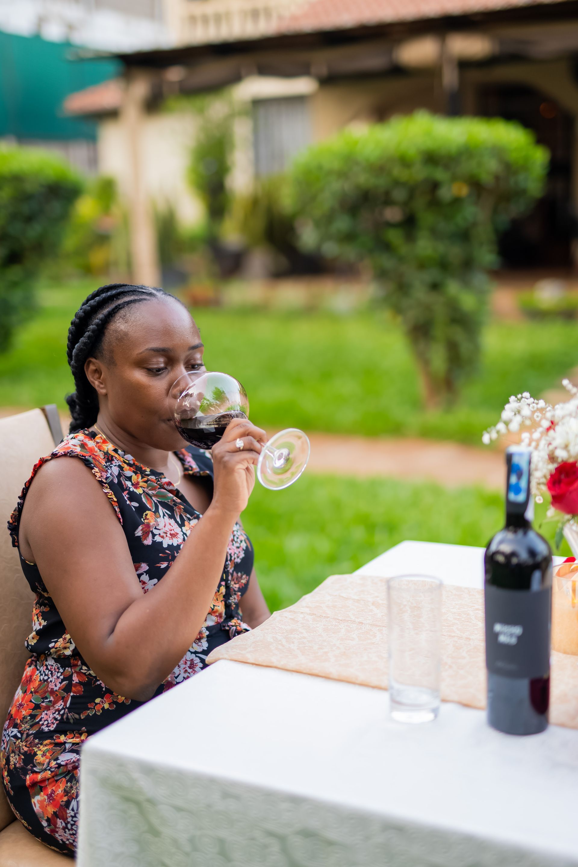 Woman with braided hair sips red wine outdoors at a table, bottle of wine and glass present.