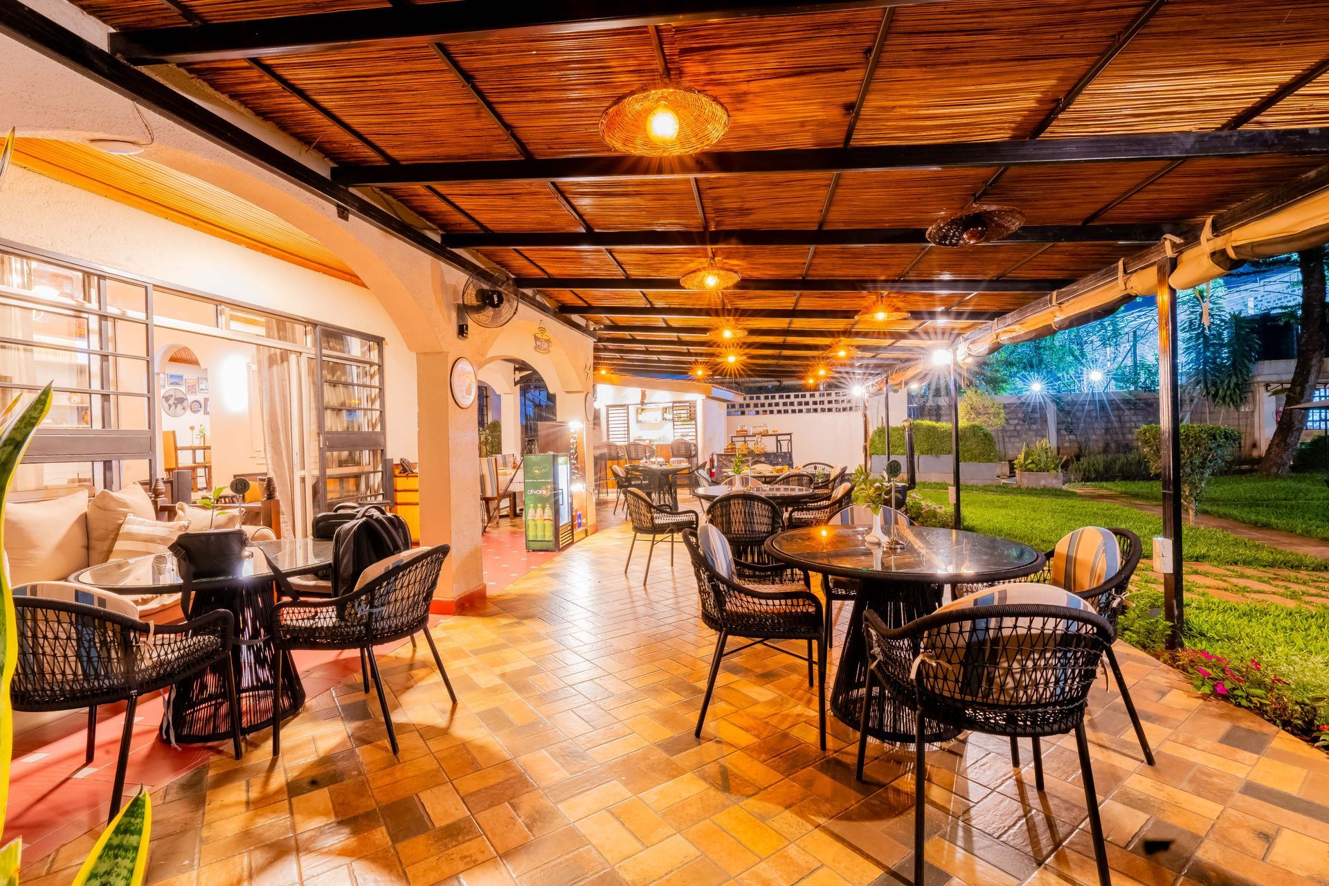 Outdoor cafe patio with tables, chairs, and decorative lighting. The space has a tiled floor and is surrounded by greenery.
