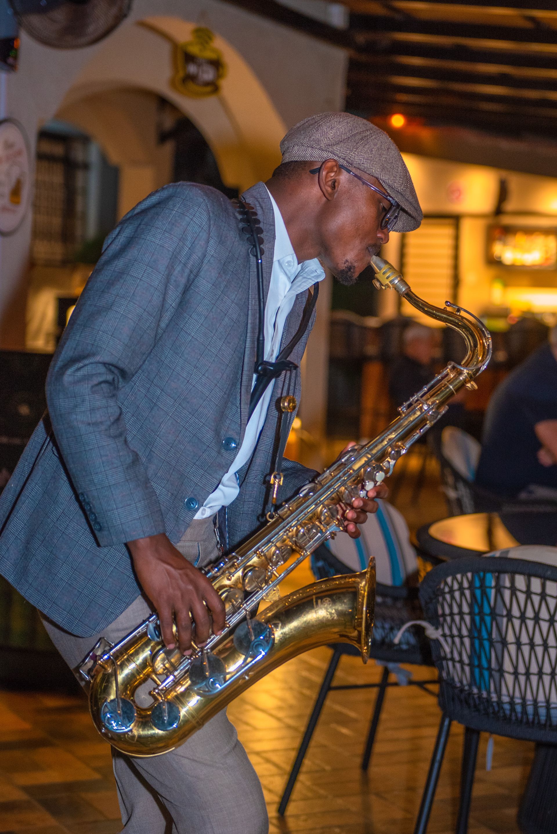 Saxophonist playing in a bar, wearing a cap and blazer. He's holding a golden saxophone playing with a focused expression.