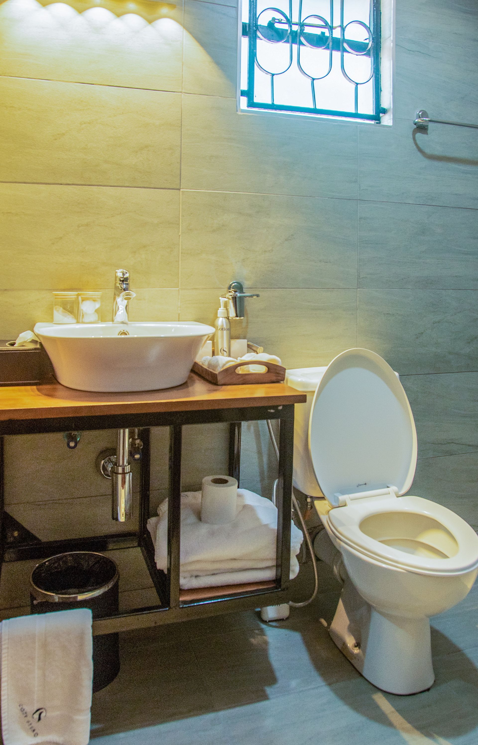 Bathroom with a sink, toilet and towels. The sink sits on a wooden counter with metal supports. A window is above the toilet.