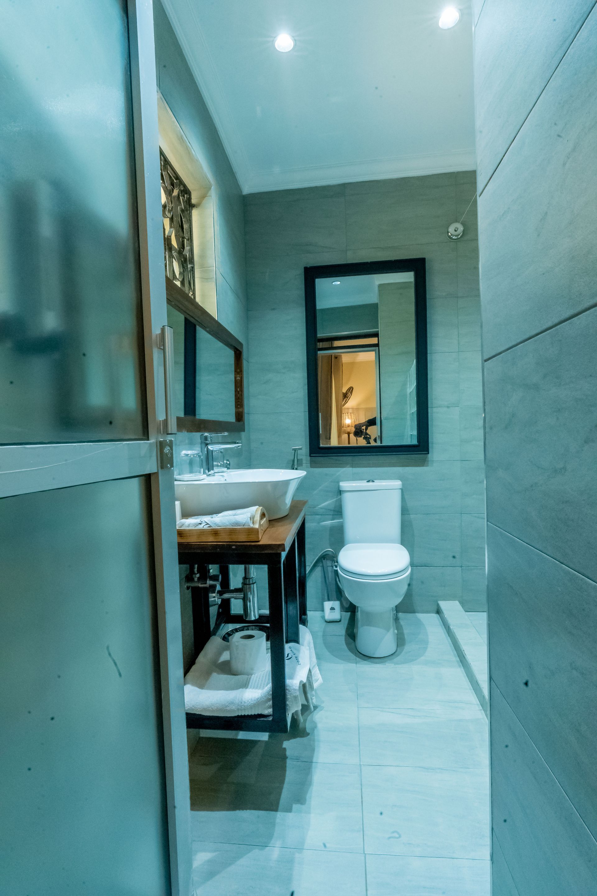 Narrow bathroom with gray tiled walls and floor, toilet, sink with mirror, and glass shower door.