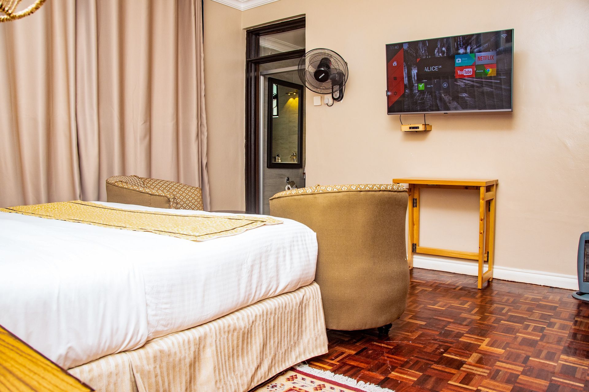 Bedroom with bed, TV, armchair, and a small wooden table on parquet floors. Beige walls and curtains.