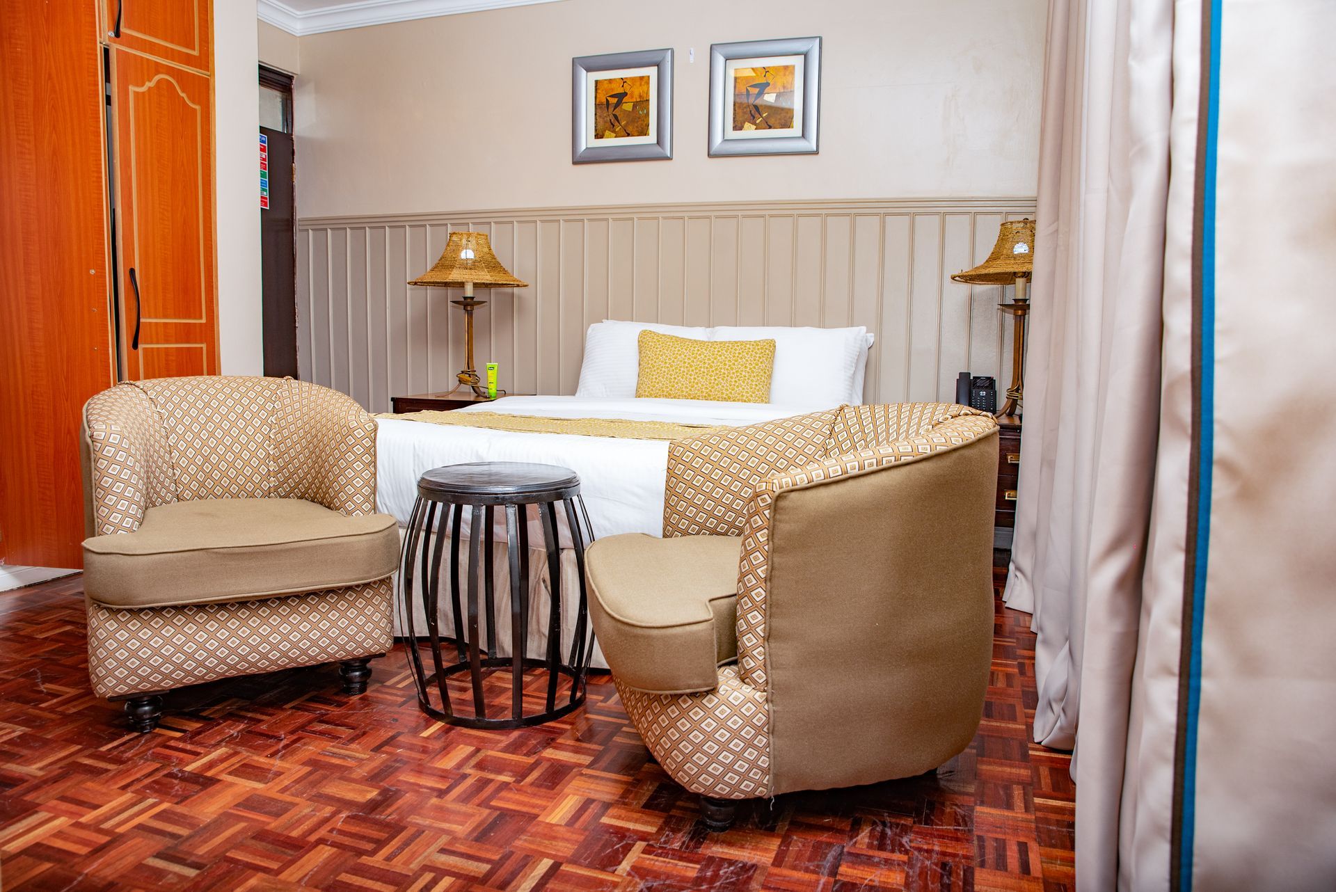 A bedroom with a bed, two patterned armchairs, a small table, and wooden flooring. Two lamps sit on either side of the bed.