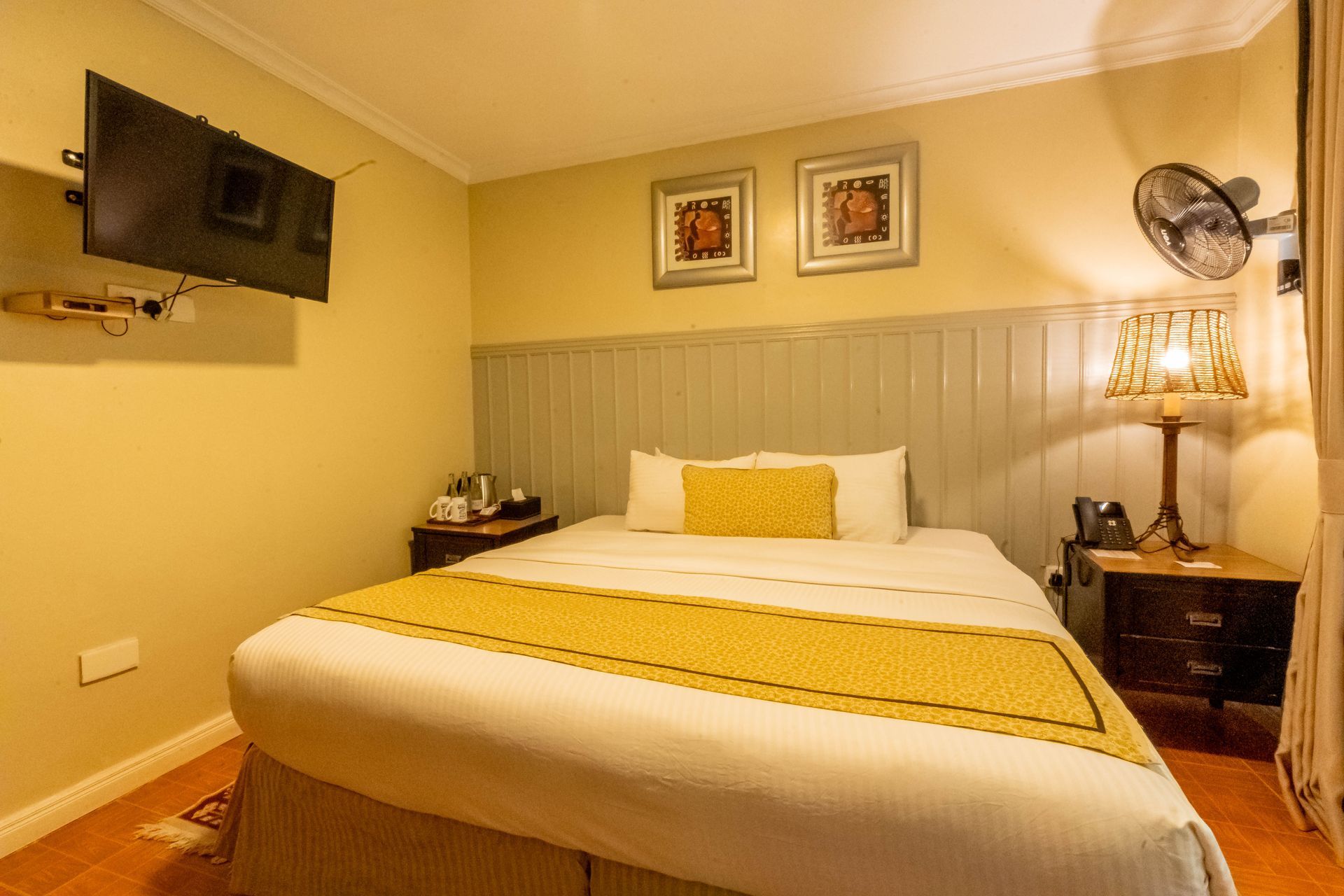 A hotel bedroom with a bed, TV and bedside table.  Yellow and cream bedding contrasts with the light wood paneling and walls.